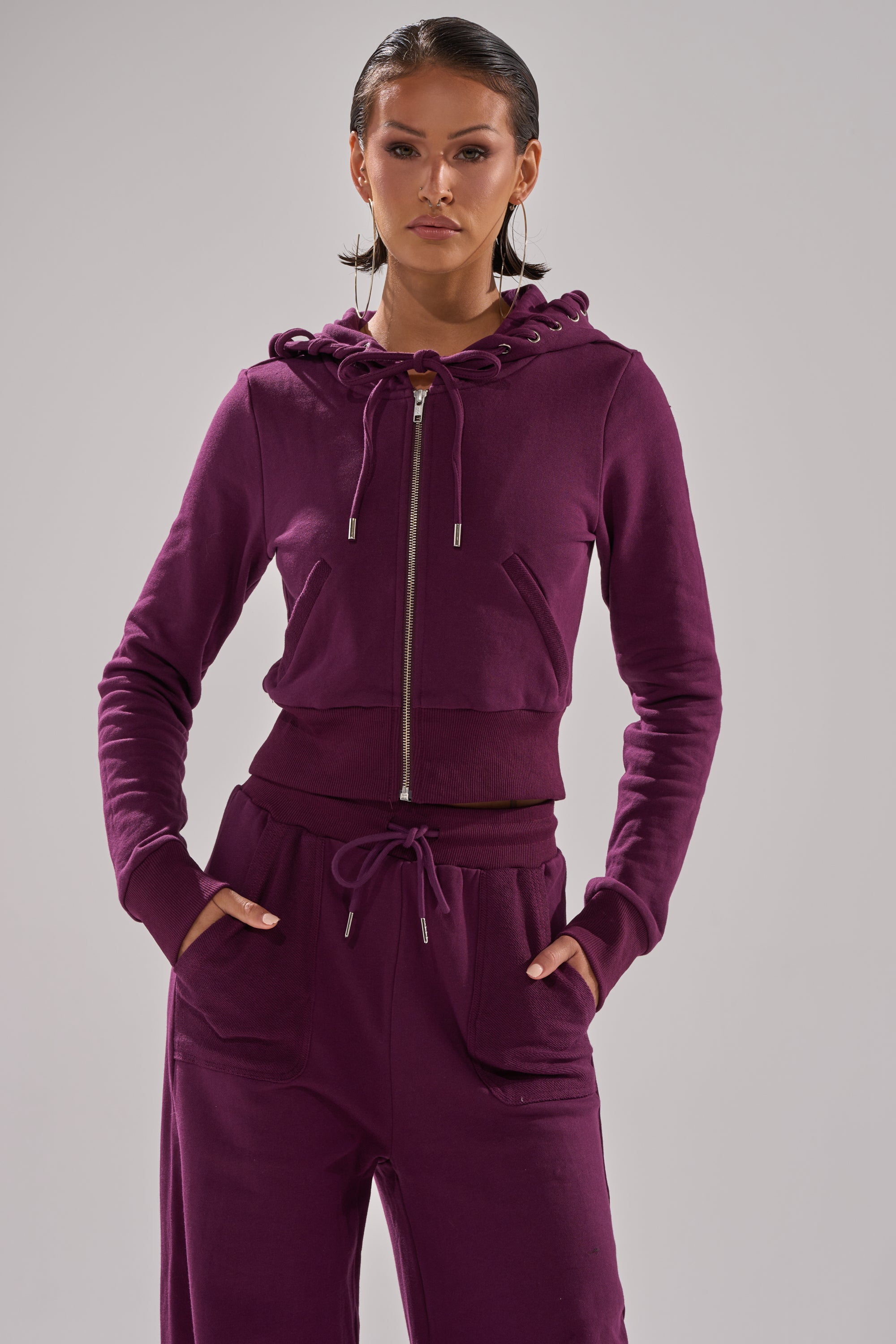 A woman stands confidently with her hands in her pockets, wearing the SHE'S GONE, YOU'RE FINE ZIP UP SWEATSHIRT in deep purple and matching pants against a plain light gray background. 4