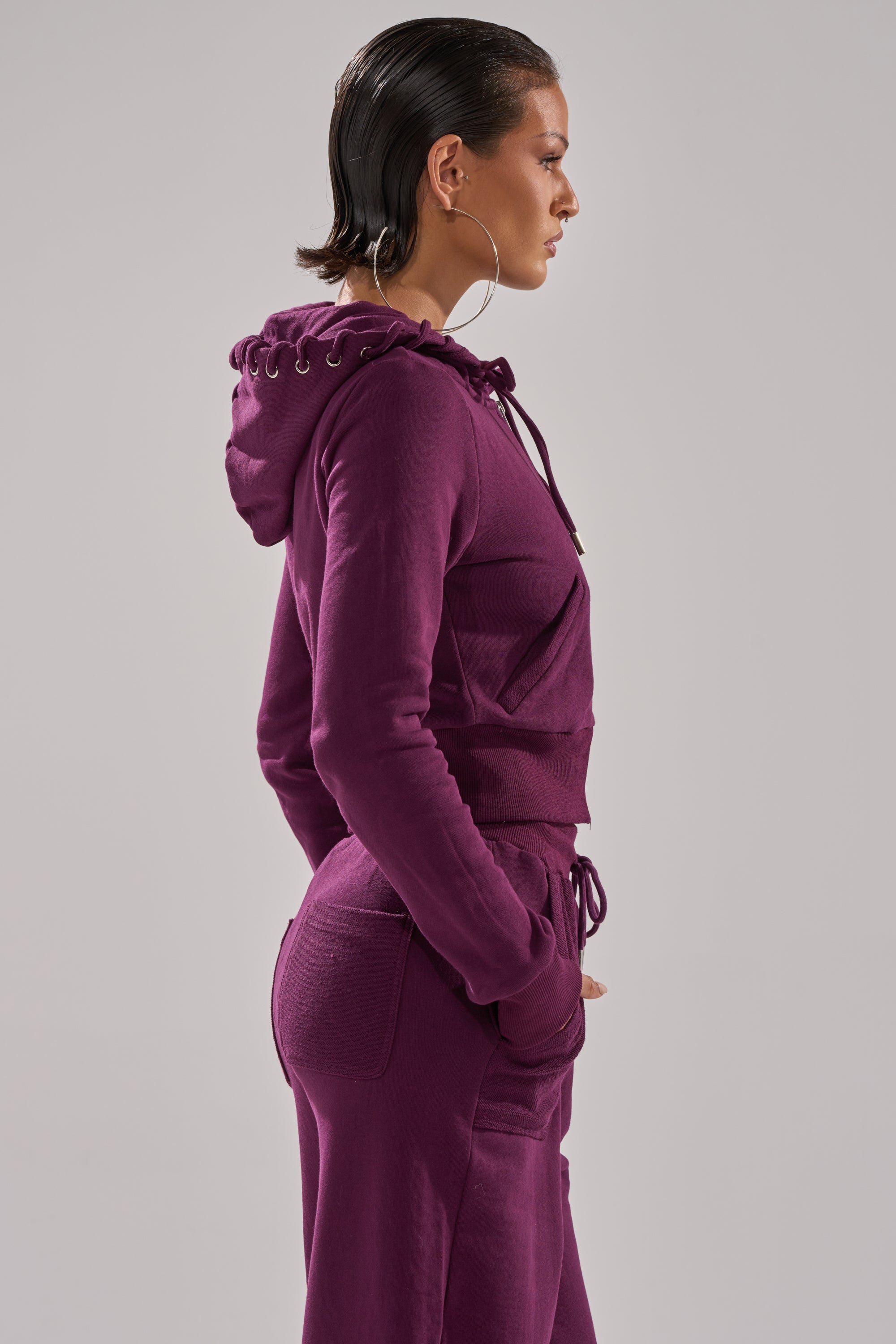 A woman with slicked-back hair and large hoop earrings wears the SHE'S GONE, YOU'RE FINE ZIP UP SWEATSHIRT and matching sweatpants, standing in profile against a plain light gray background. 5