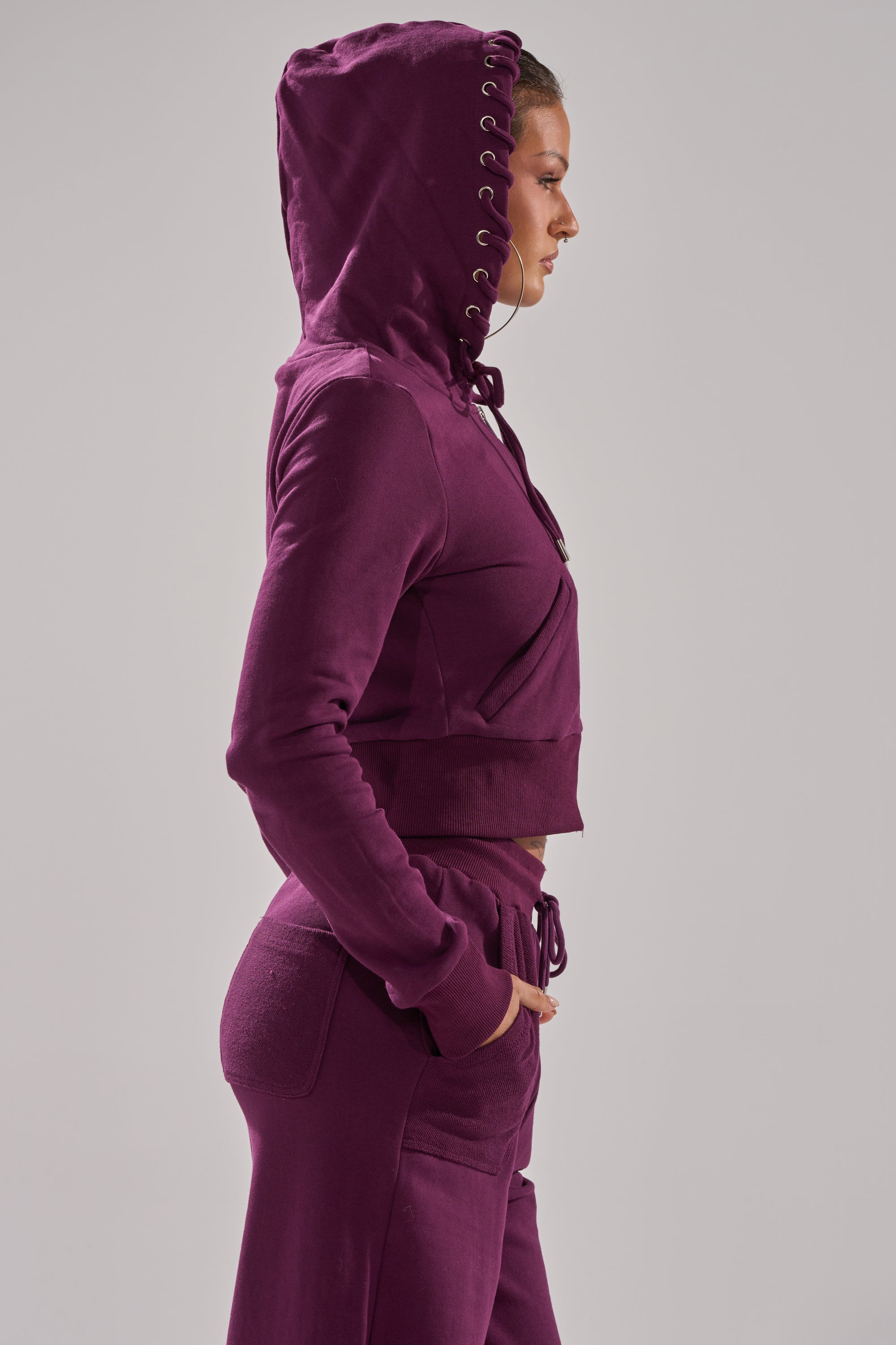 A woman in side profile wears a matching deep purple SHE'S GONE, YOU'RE FINE ZIP UP SWEATSHIRT with a lace-up hood and jogger pants against a plain light gray background. 6