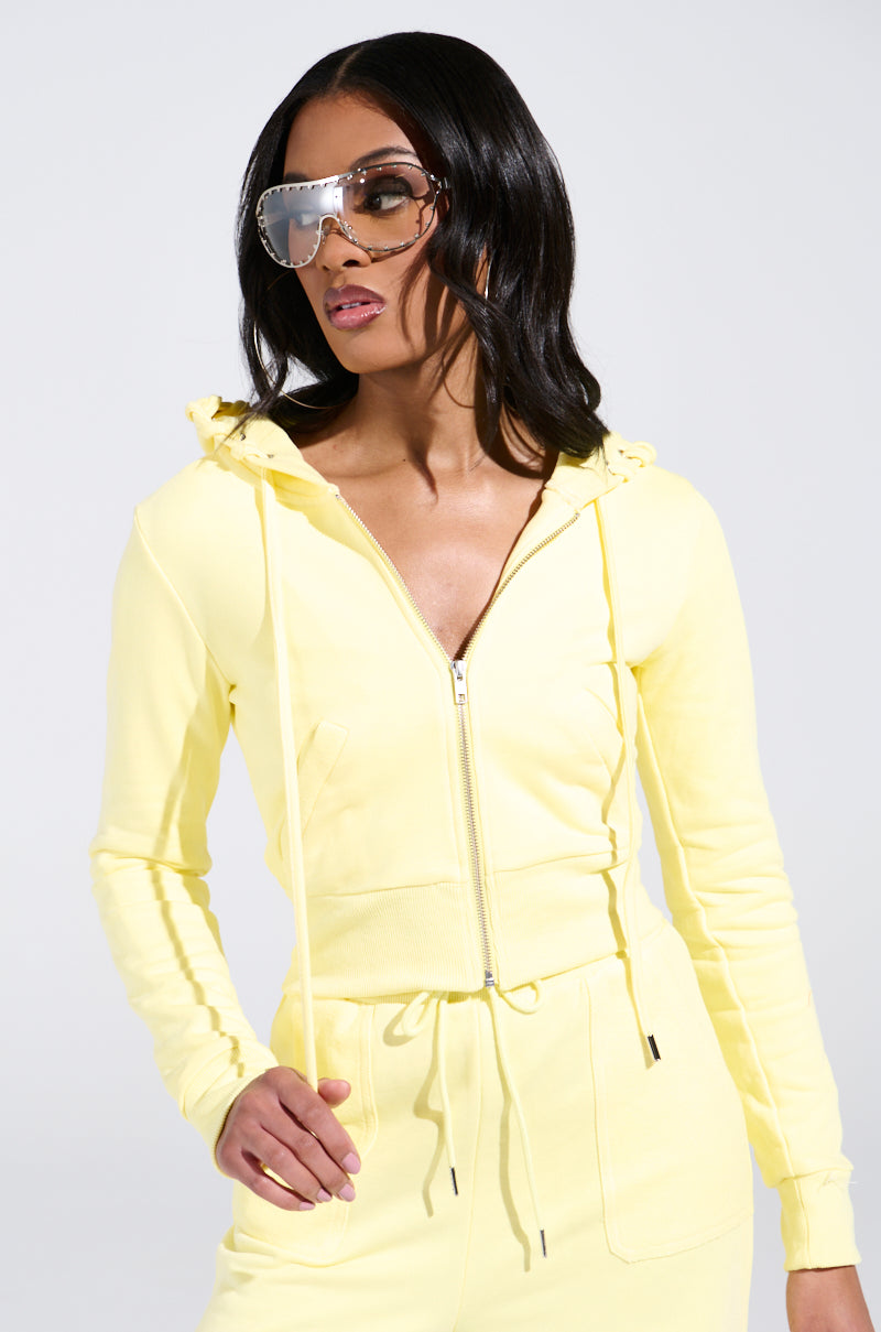 Someone confidently flaunts a monochromatic style in an eye-catching BUTTERFINGER SWEATSHIRT paired with matching pants featuring a front zipper. Their long dark hair flows under oversized reflective sunglasses, completing the bold AKIRA Label look against a plain, light background.