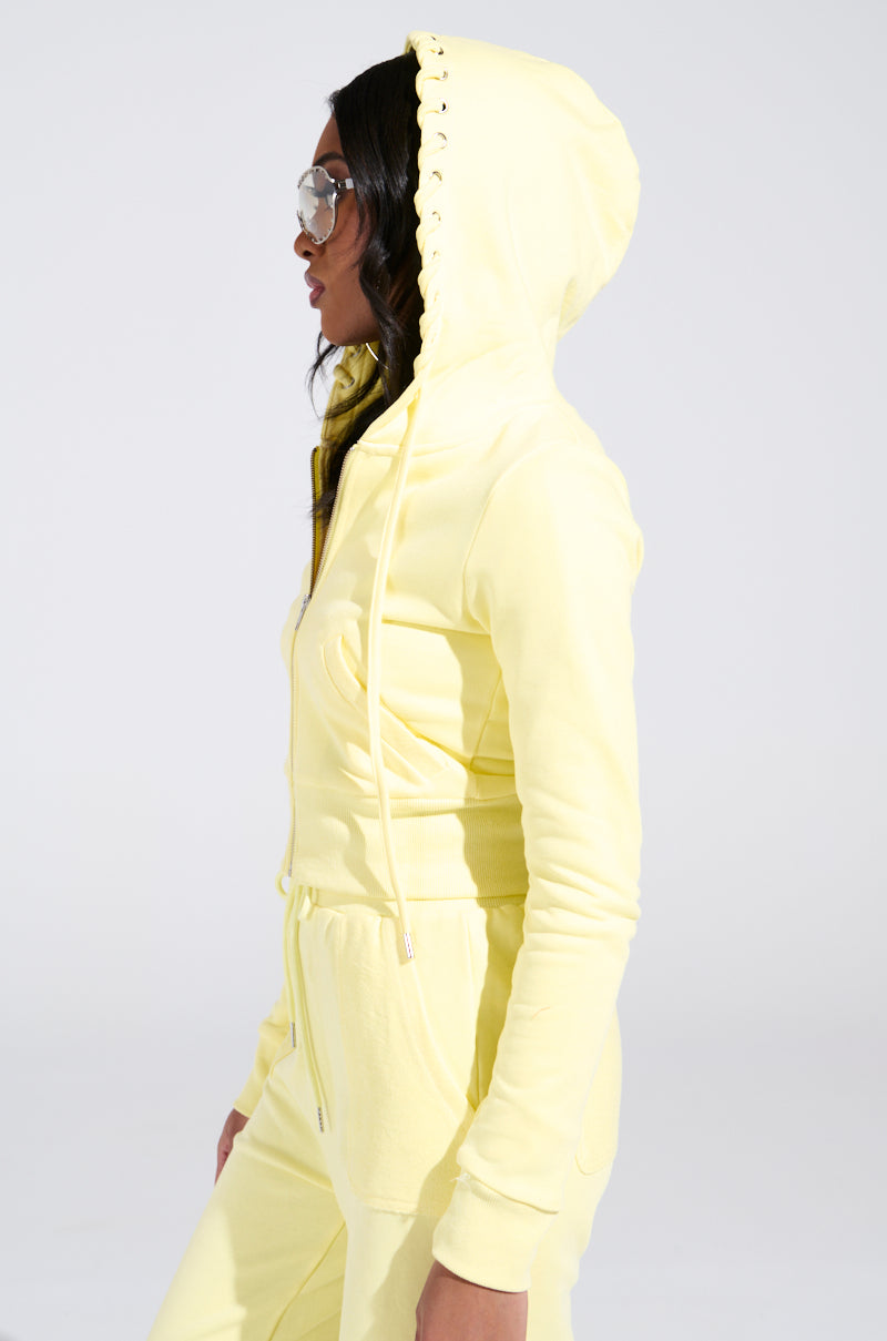 Wearing the BUTTERFINGER SWEATSHIRT, a person in a yellow hooded tracksuit and sunglasses poses against a plain background, embodying an edgy monochromatic look and creating a striking visual statement.