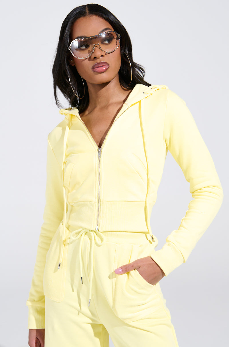 A person confidently poses in a yellow BUTTERFINGER SWEATSHIRT and matching pants, accessorized with large sunglasses and hoop earrings. The plain, light background highlights their striking AKIRA Label monochromatic look.
