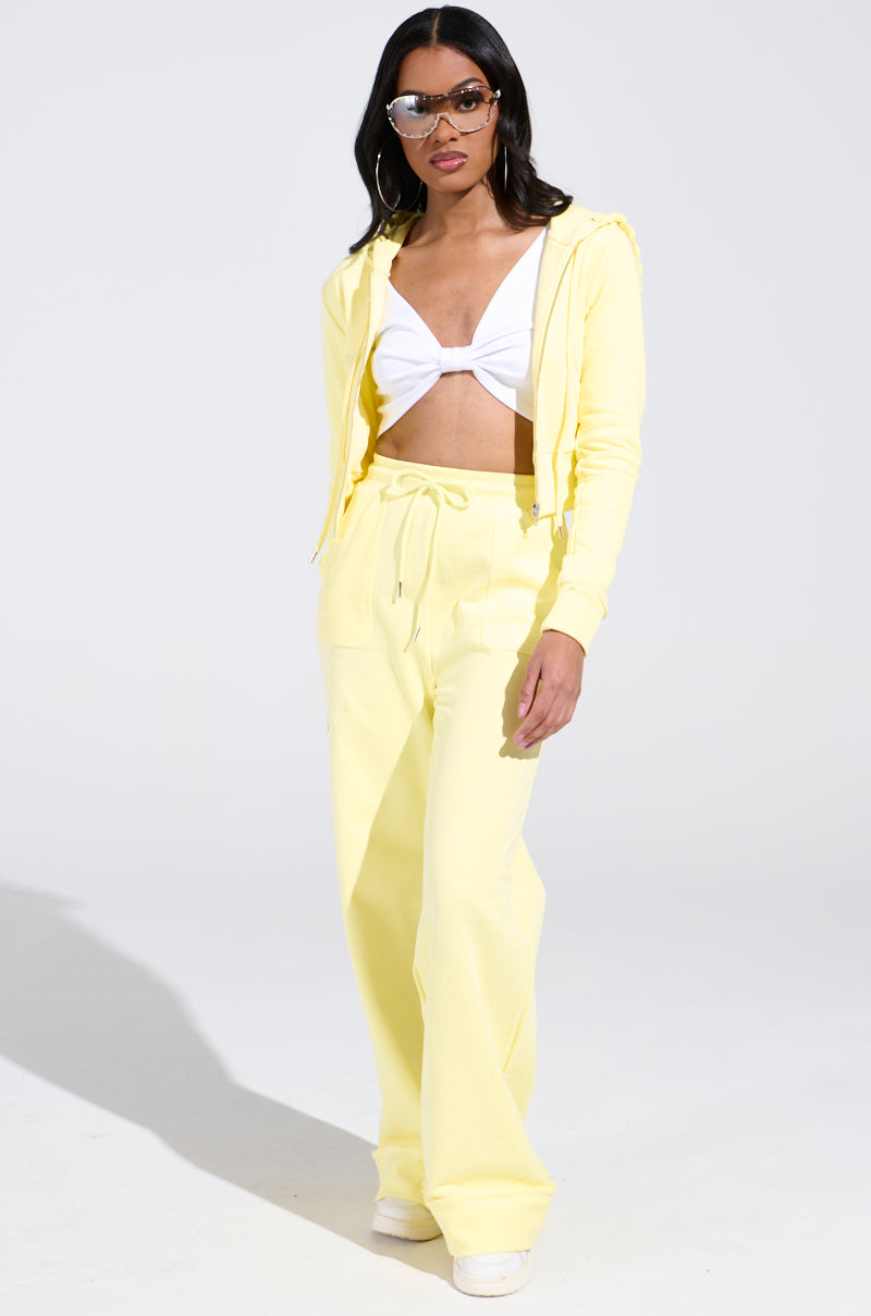 A person in sunglasses and hoop earrings wears a BUTTERFINGER SWEATSHIRT with wide-legged pants for a monochromatic look. A white tied crop top is visible underneath as they stand against a plain white background.