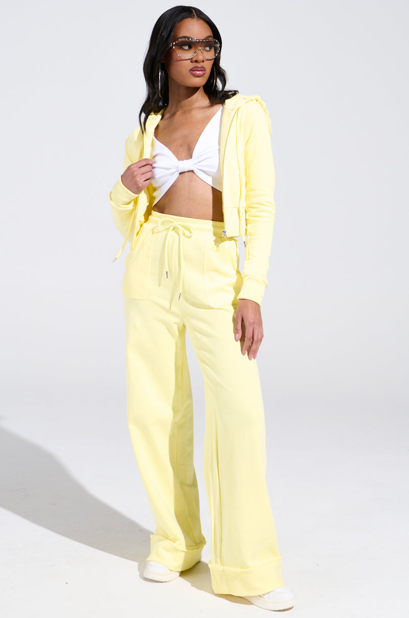 In a studio, a woman showcases her monochromatic look with a yellow BUTTERFINGER SWEATSHIRT layered over a white top. Her long, dark hair complements her chic sunglasses as she makes a stylish statement with AKIRA Label hoodie and wide-legged pants.