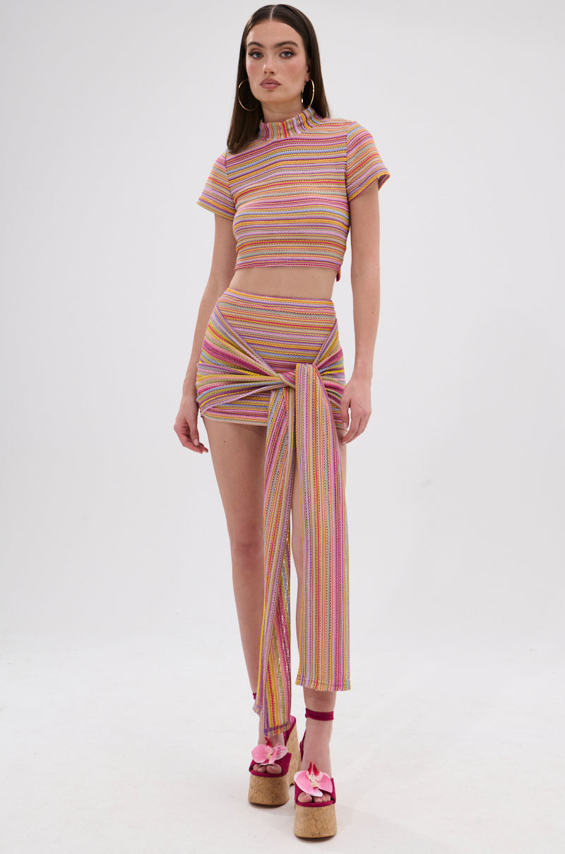 A woman with long brown hair wears the MORNING SHINE CROCHET MINI SKIRT and matching colorful, striped crop top. She styles the look with hoop earrings and pink-bow platform sandals, posed in a plain white studio. 0