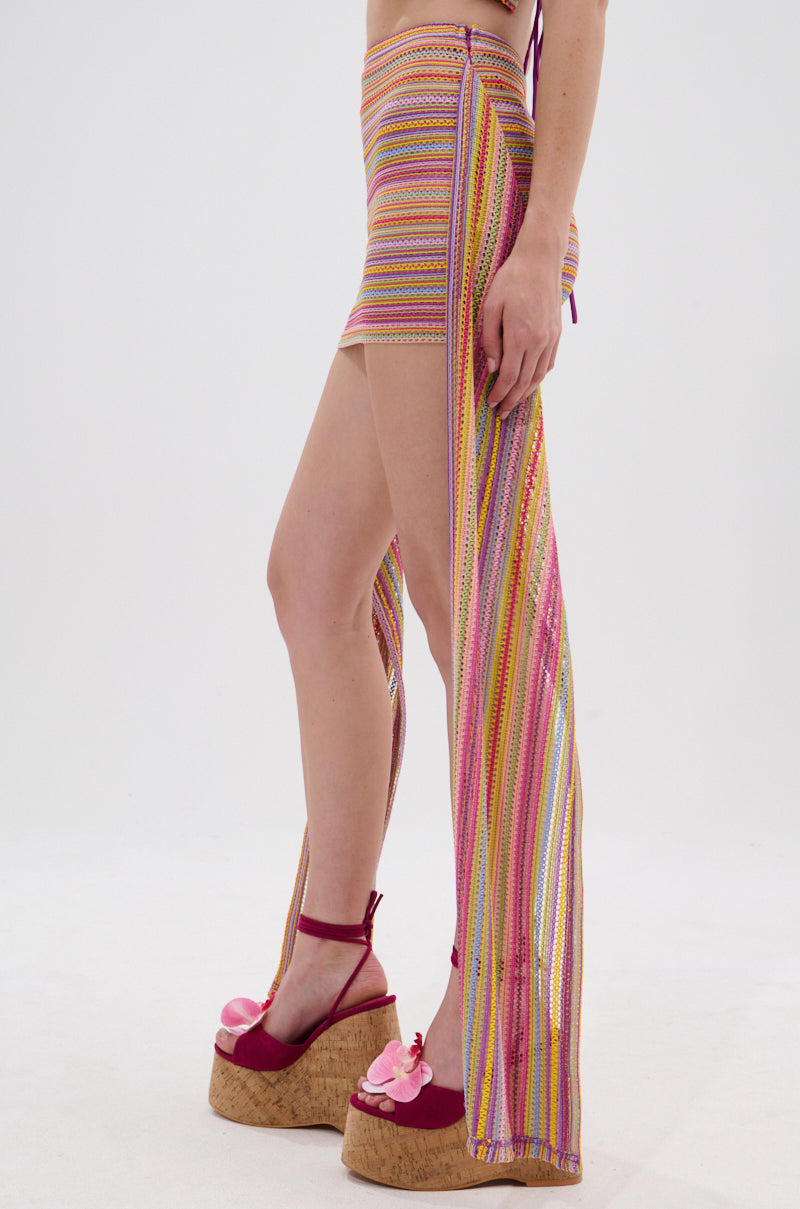 Model wears the MORNING SHINE CROCHET MINI SKIRT—colorful and striped with an asymmetrical cut—and pink platform sandals with floral accents, standing against a plain white background. 3