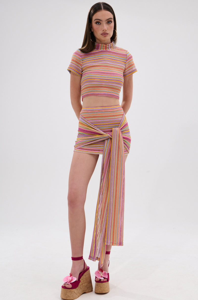 A woman poses against a white background wearing the MORNING SHINE CROCHET MINI SKIRT, a colorful striped two-piece with a short-sleeve crop top, mini skirt with draped sash, and pink platform sandals. 7