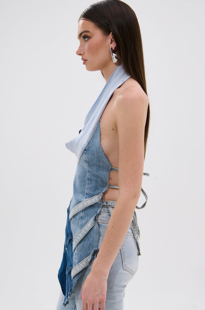 A woman with long straight brown hair models the PATCHED UP COWL NECK DENIM HALTER TOP, featuring patchwork denim, lace-up strappy details, and exposed sides. She pairs it with light-wash jeans and large silver earrings against a plain white background. 2