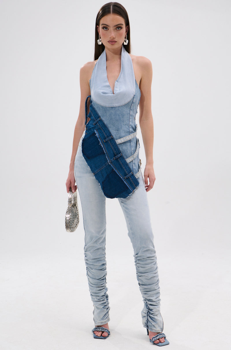 Against a plain background, a woman models Y2K style in the PATCHED UP COWL NECK DENIM HALTER TOP with matching ruched denim pants, silver clutch, dark blue belt bag, heeled sandals, and hoop earrings. 6