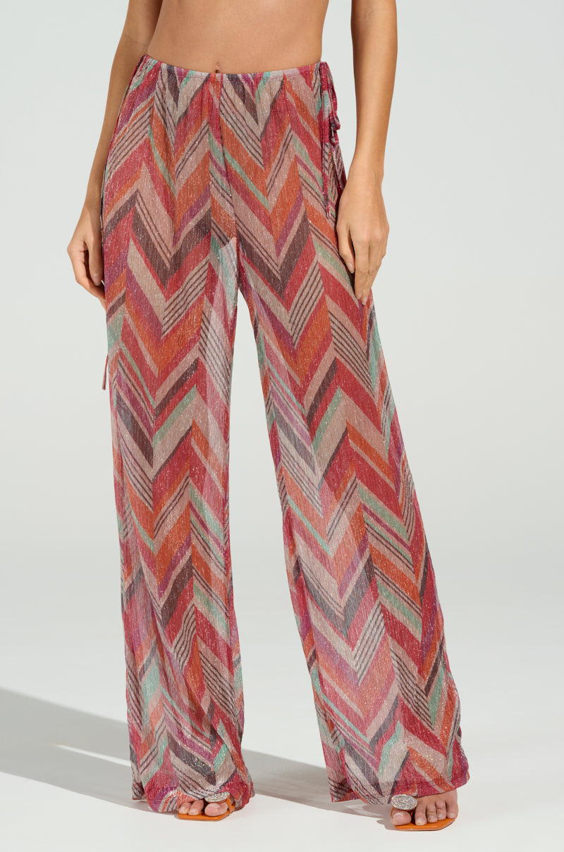Barefoot, a person models CALI FAYE WIDE LEG PANT with a vivid chevron print in red, orange, pink, and green. The pants are designed with an elasticized waistband for comfort against a plain light background.
