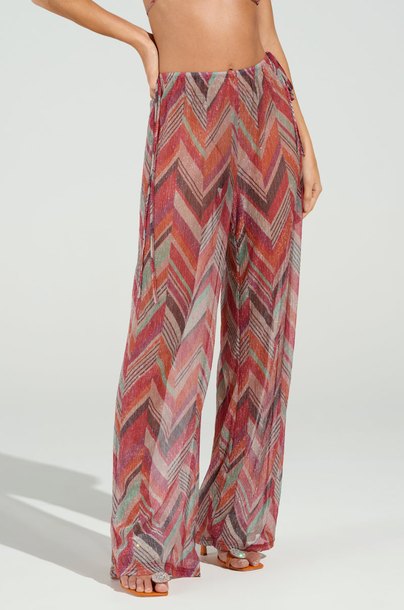 A person confidently stands on a light background wearing CALI FAYE WIDE LEG PANT. These colorful, sheer beach pants showcase a chevron print in vibrant red, pink, and green hues. Featuring an elasticized waistband and a wide-leg design, they place their right hand on their hip.