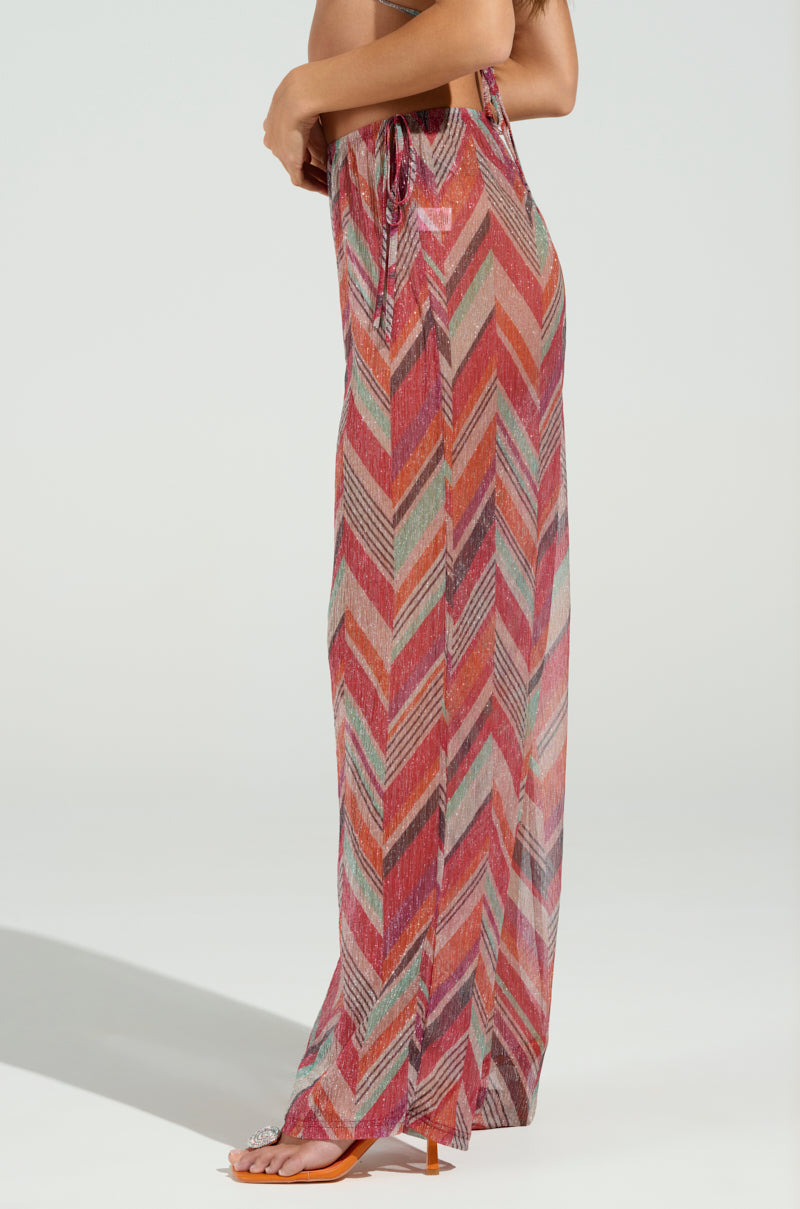A person is wearing colorful chevron-patterned CALI FAYE WIDE LEG PANTS featuring an elasticized waistband and a high slit on the side. The lightweight fabric flows elegantly, matched with open-toed tan sandals, all set against a plain light background.