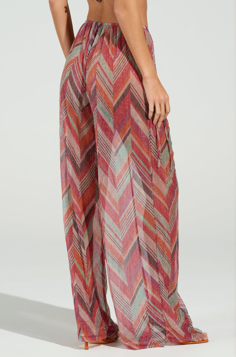 A person models CALI FAYE WIDE LEG PANT with a colorful chevron print and an elastic waistband against a plain background. The pants display vibrant shades of red, pink, and teal as the individual places their hand on their hip.