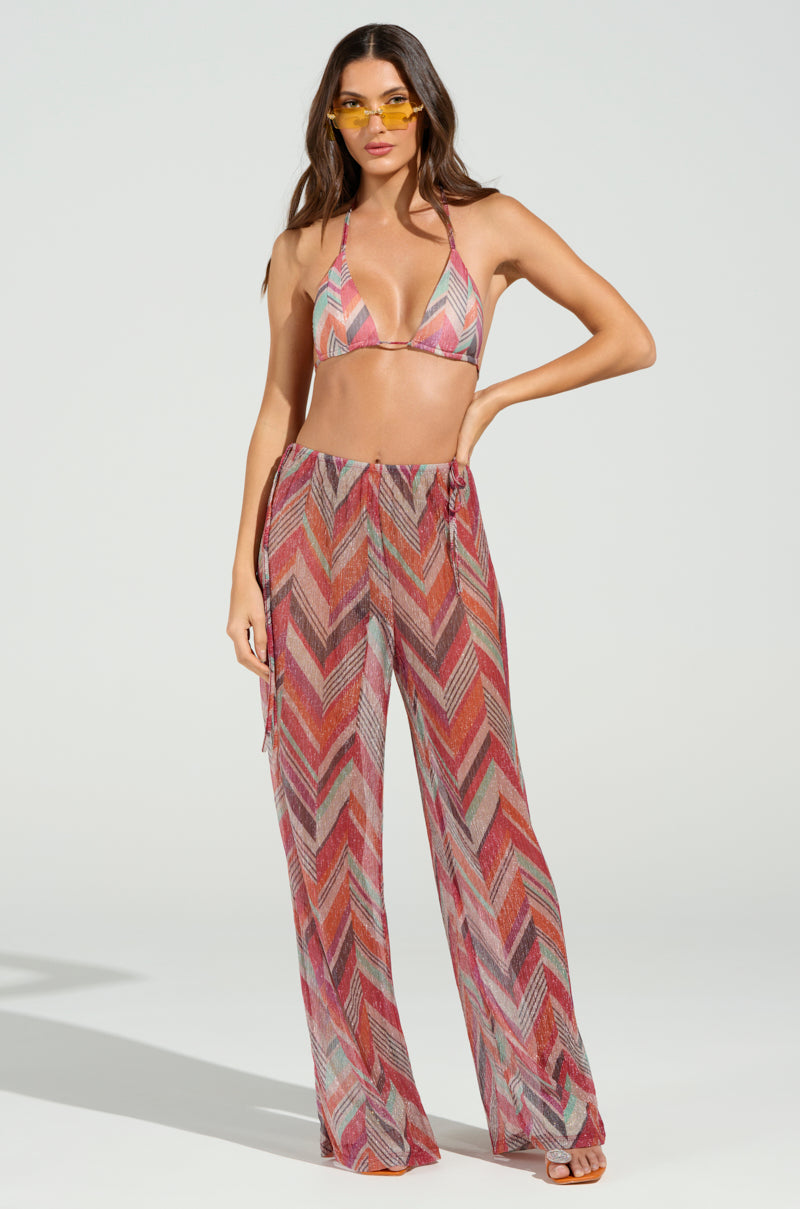 Wearing a CALI FAYE WIDE LEG PANT paired with a vibrant chevron bikini top and yellow sunglasses, the person stands against a neutral backdrop. Their long hair flows gently in the breeze.