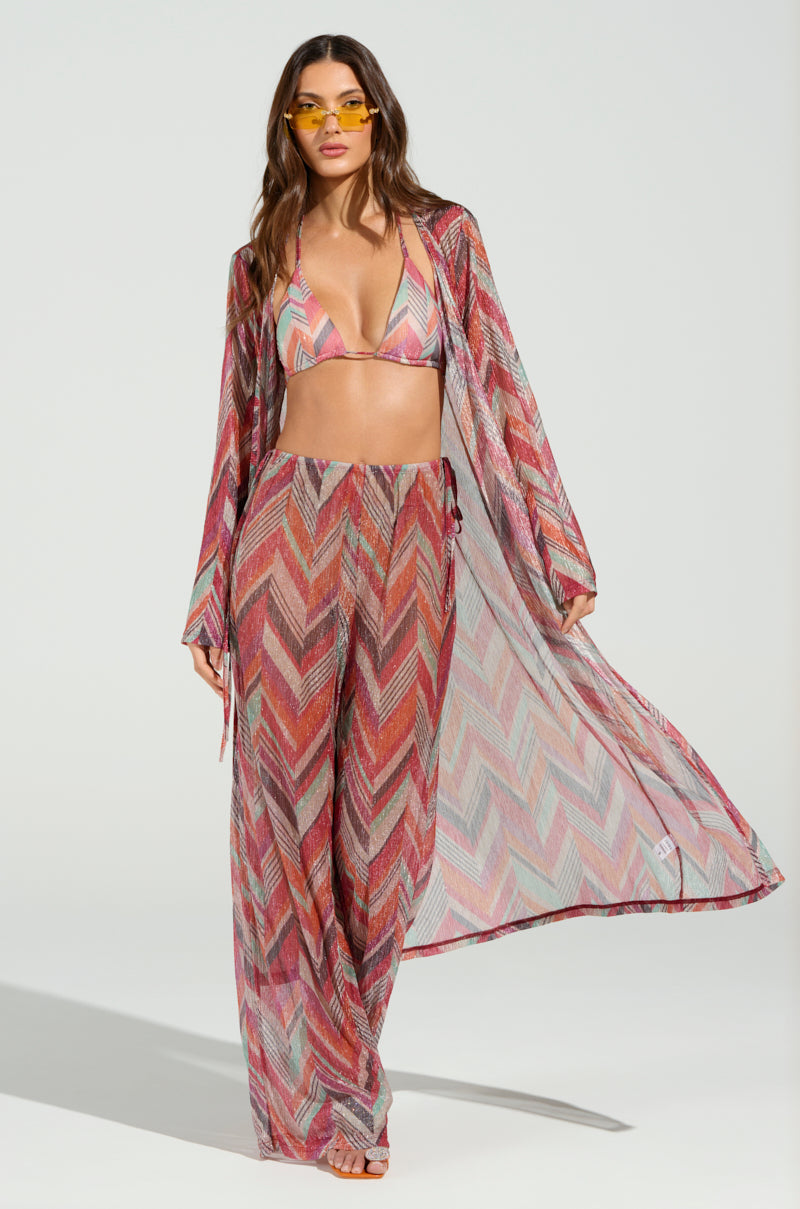 A woman models the CALI FAYE WIDE LEG PANT with a vibrant, chevron-patterned bikini top against a plain background. She wears sunglasses, her long hair cascades down, and she is draped in a flowing cover-up.