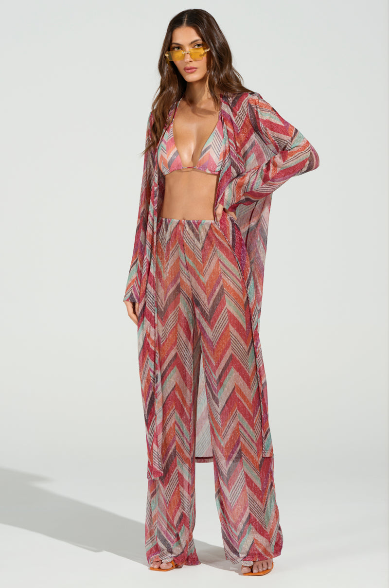 Wearing the CALI FAYE WIDE LEG PANT in a vibrant chevron-print sheer design, she pairs it with a matching bikini top and open cardigan. Her look is completed with yellow-tinted sunglasses, while her long hair flows freely against a simple backdrop.