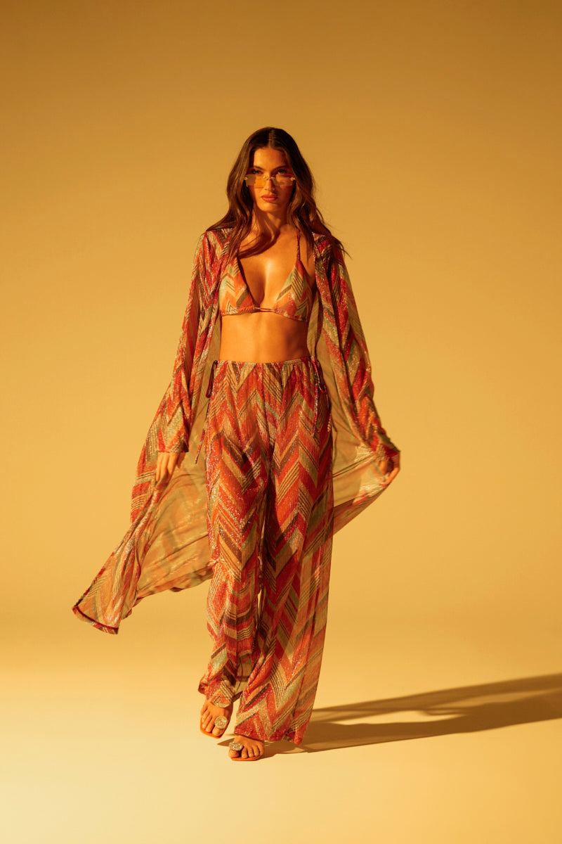 Confident in a chevron print outfit, a person shines wearing the CALI FAYE LONGLINE DUSTER and sunglasses. The ensemble flows gracefully against a sunlit, golden beach backdrop.