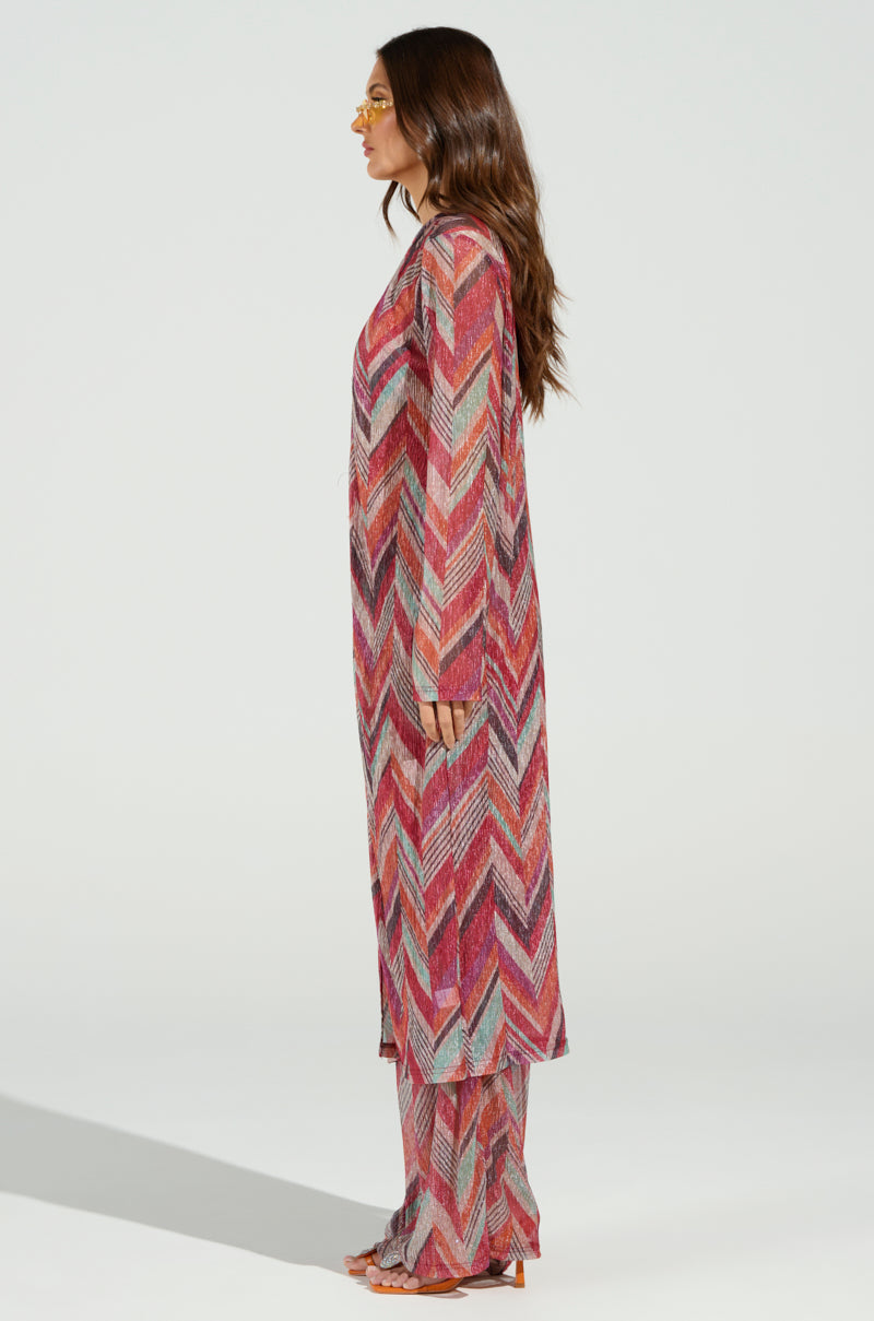 A woman poses sideways in a CALI FAYE LONGLINE DUSTER, featuring a vibrant geometric chevron print in red, pink, and gray. Her long wavy brown hair pairs perfectly with her orange-tinted sunglasses, exuding an effortlessly chic beachy vibe against the plain white backdrop.