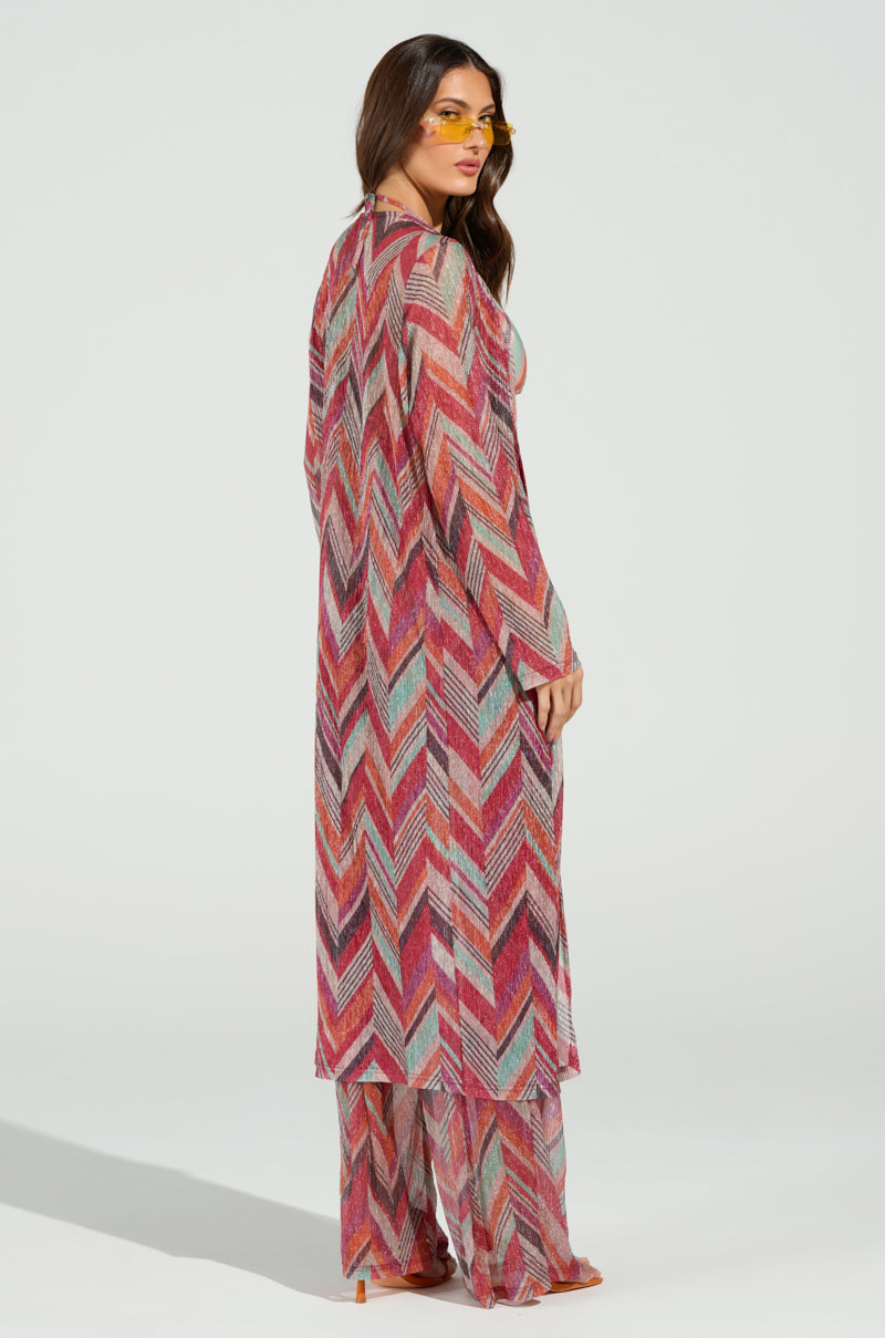A person in a vibrant CALI FAYE LONGLINE DUSTER glances over their shoulder, with long hair, yellow-tinted sunglasses, and matching wide-leg pants against a plain white backdrop.