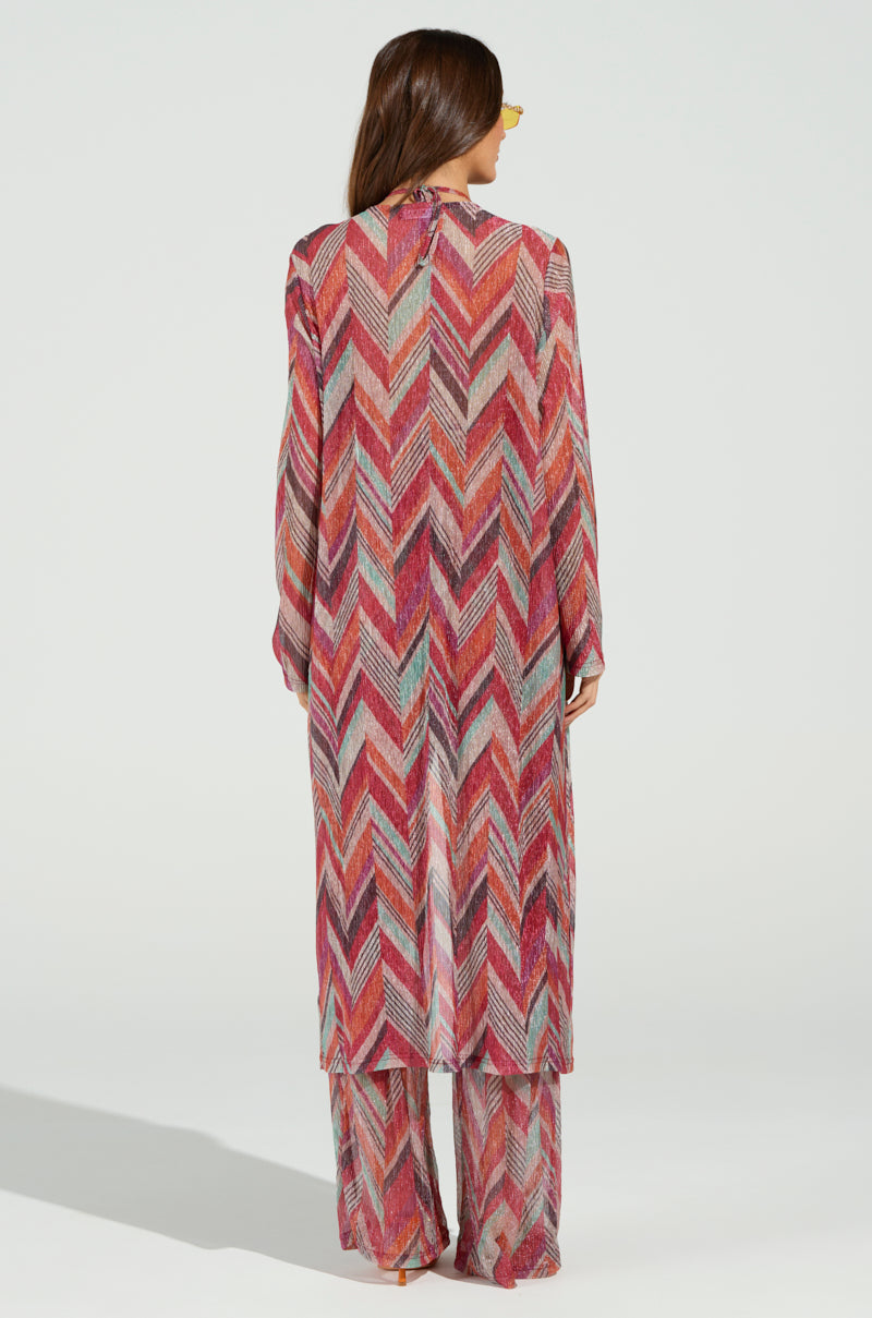 A person with long hair stands facing away, wearing a colorful maxi dress featuring a chevron print in shades of pink, red, gray, and orange. Beneath the dress peeks the CALI FAYE LONGLINE DUSTER, paired with beachy wide-leg pants for a complete look.