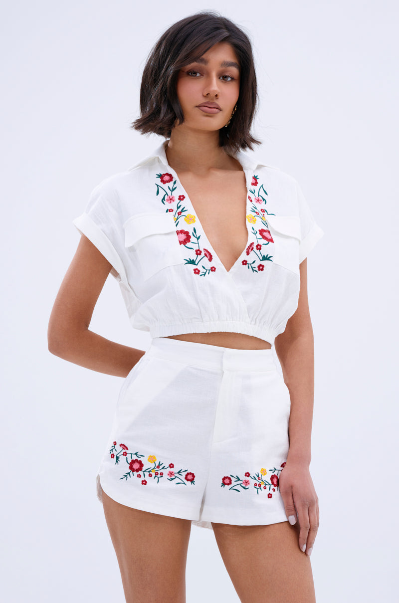 A woman wears the DAY TIME VIBES BUTTON DOWN TOP, a white cropped blouse with colorful floral embroidery at the neckline, paired with high-waisted shorts for a fresh, stylish look against a light background.