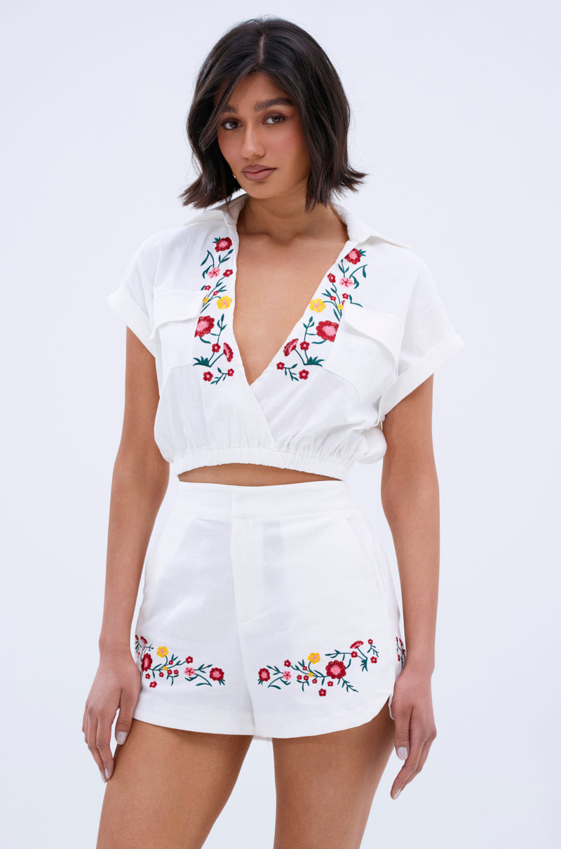 A woman wears the DAY TIME VIBES BUTTON DOWN TOP, a white cropped blouse with colorful floral embroidery and matching high-waisted shorts, standing against a plain light background and looking at the camera.