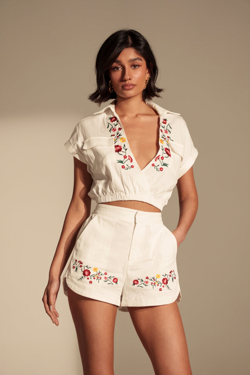 A woman stands against a neutral background, wearing the DAY TIME VIBES BUTTON DOWN TOP—a white cropped blouse with colorful floral embroidery—paired with matching high-waisted shorts.