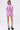 A woman with short dark hair, glasses, and black platform heels stands against a plain background in the AKIRA Label TAKE ME TO IBIZA TIE FRONT TOP, a pink and white striped blouse with ruffles, paired with matching high-waisted shorts.