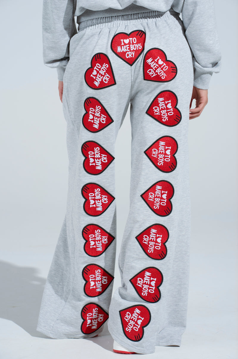 A person sported I LOVE TO MAKE BOYS CRY WIDE LEG SWEATPANT, featuring a high-rise fit in gray with red heart patches and "I ❤️ TO MAKE BOYS CRY" in white text cascading down the legs.