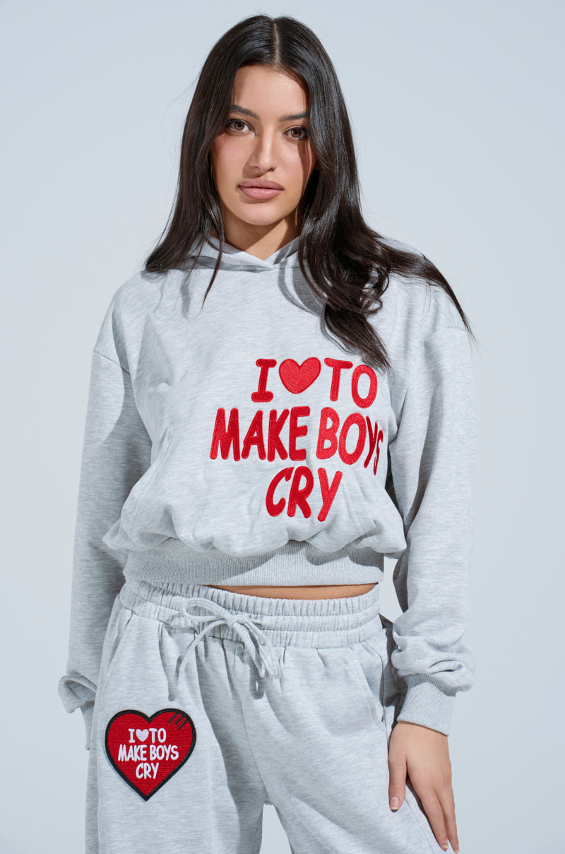 A person with long dark hair wears an oversized gray I LOVE TO MAKE BOYS CRY HOODED SWEATSHIRT and matching sweatpants featuring "I ♥ TO MAKE BOYS CRY" text. The backdrop is a simple, light hue.