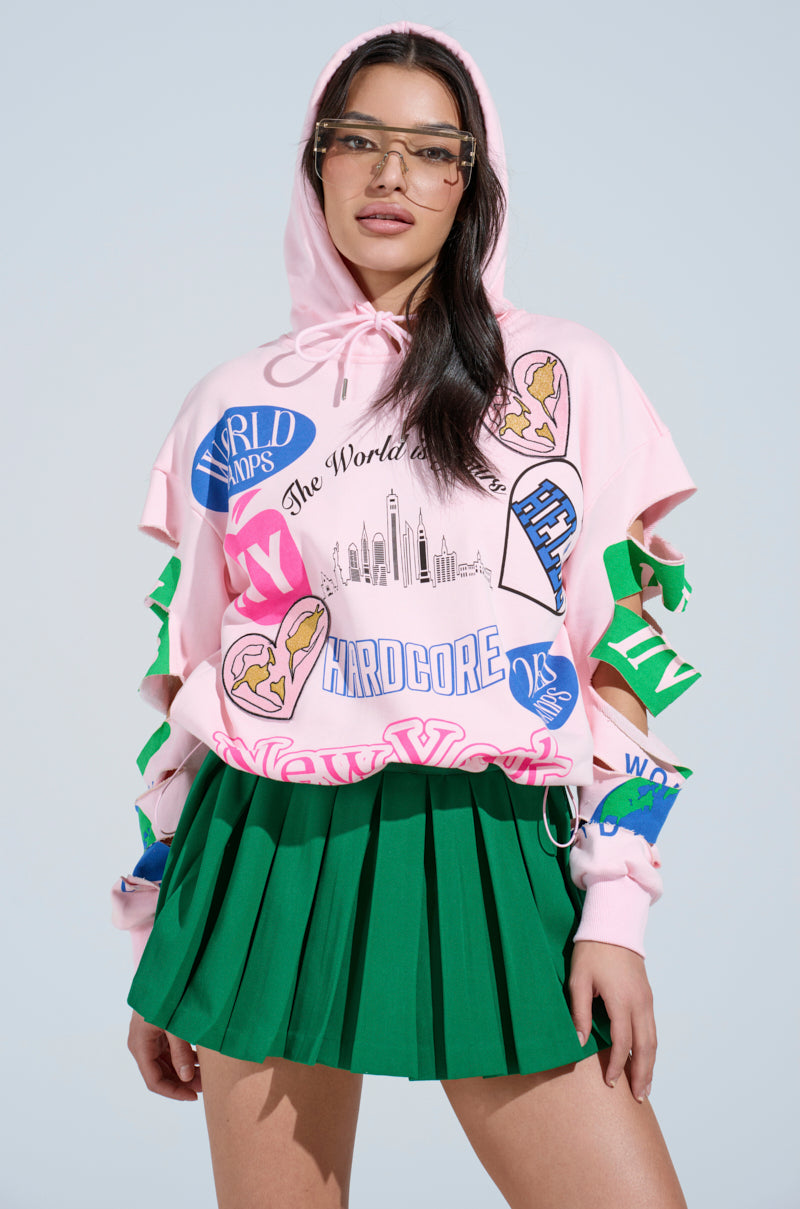 Against a light blue background, a woman in an oversized pink NEW YORK BABY SWEATSHIRT with patches reading "World" and "Hardcore" pairs it with glasses and a green pleated skirt.