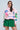 Against a light blue background, a woman in an oversized pink NEW YORK BABY SWEATSHIRT with patches reading "World" and "Hardcore" pairs it with glasses and a green pleated skirt.