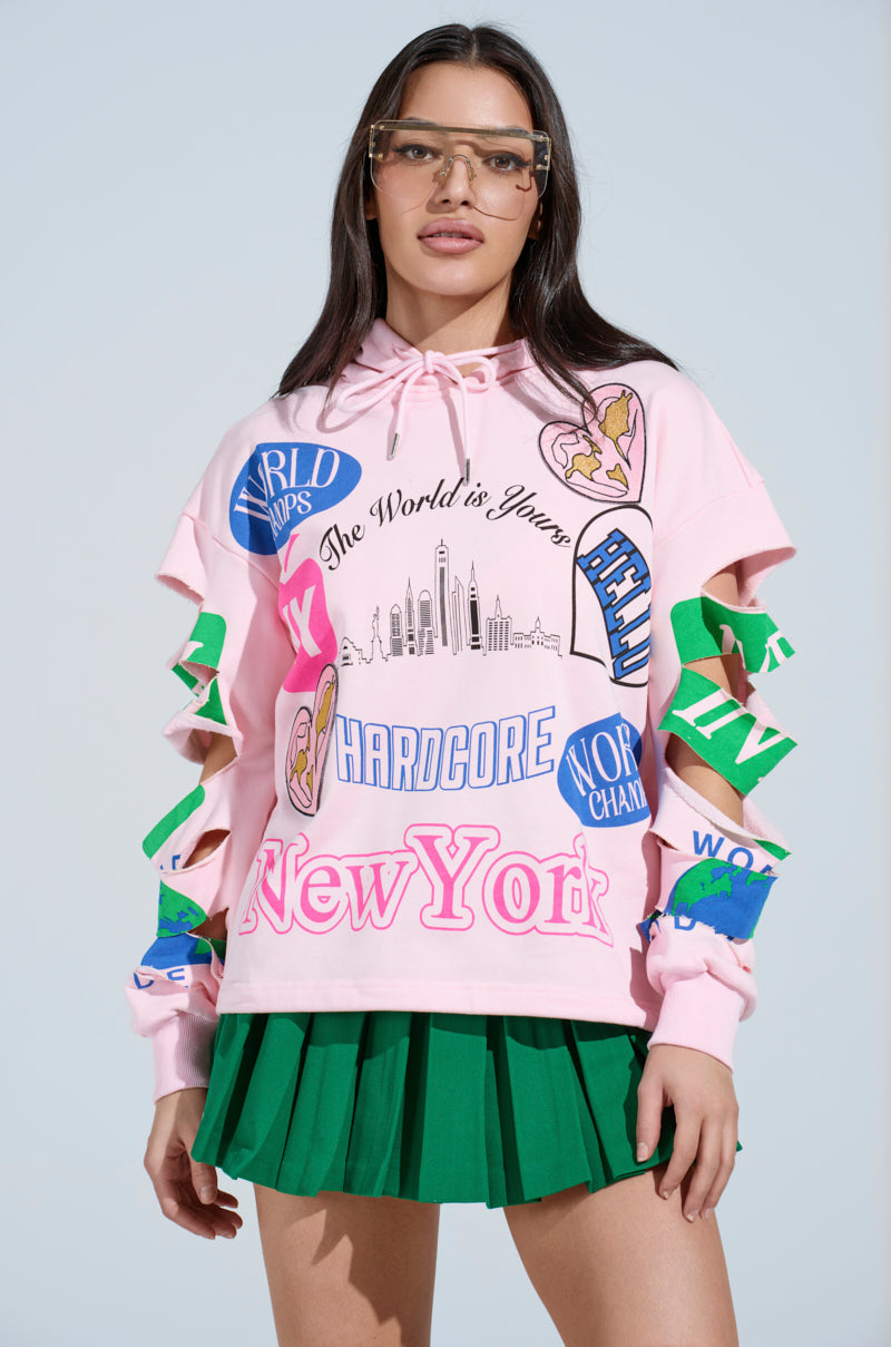 A person in glasses and a NEW YORK BABY SWEATSHIRT, an oversized pink hoodie with colorful text and graphics like "New York" and "The World is Yours," paired with a green pleated skirt, stands against a light blue background.