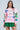 A person in glasses and a NEW YORK BABY SWEATSHIRT, an oversized pink hoodie with colorful text and graphics like "New York" and "The World is Yours," paired with a green pleated skirt, stands against a light blue background.