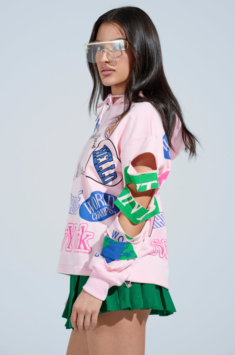 With long dark hair, the person wears large transparent sunglasses and a NEW YORK BABY SWEATSHIRT in pink with colorful patches and cut-out sleeves. Paired with a green pleated skirt, this ensemble stands out against a plain, light background.