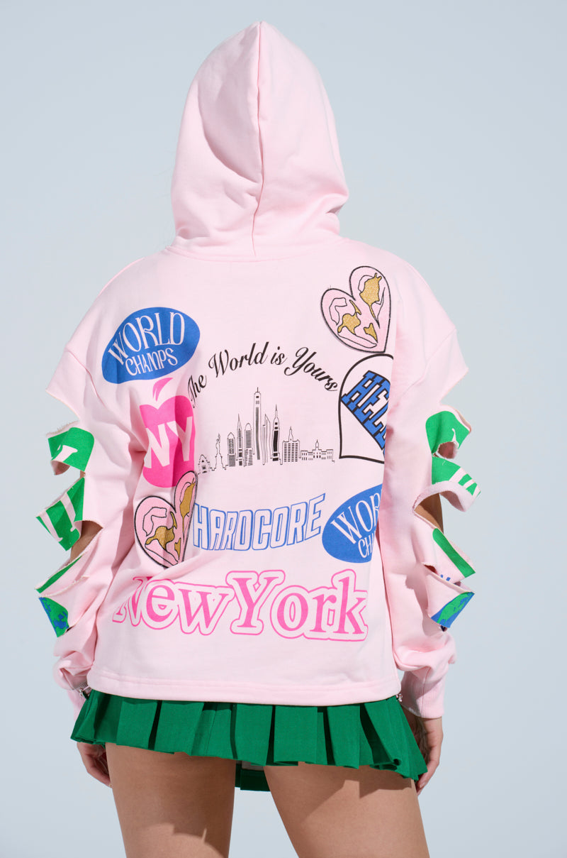 Someone is wearing an oversized pink NEW YORK BABY SWEATSHIRT adorned with patches and prints such as "World Champs," "The World is Yours," and "New York." The sweatshirt, featuring sleeve cut-outs, pairs perfectly with a green pleated skirt.