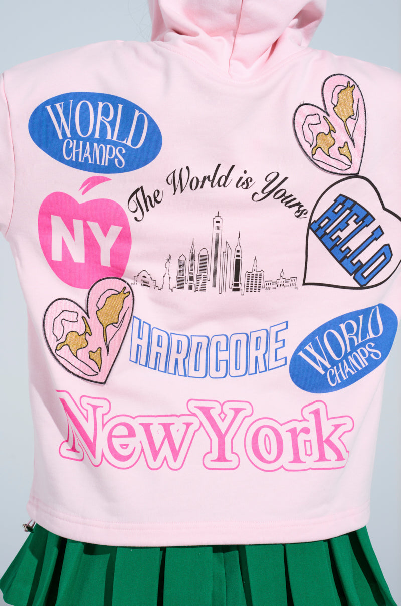 The NEW YORK BABY SWEATSHIRT, a pink cotton-blend hoodie, features "New York," "The World is Yours," a city skyline, hearts, and "World Champs." Paired with a pleated green skirt, it creates a chic urban vibe with its oversized design.