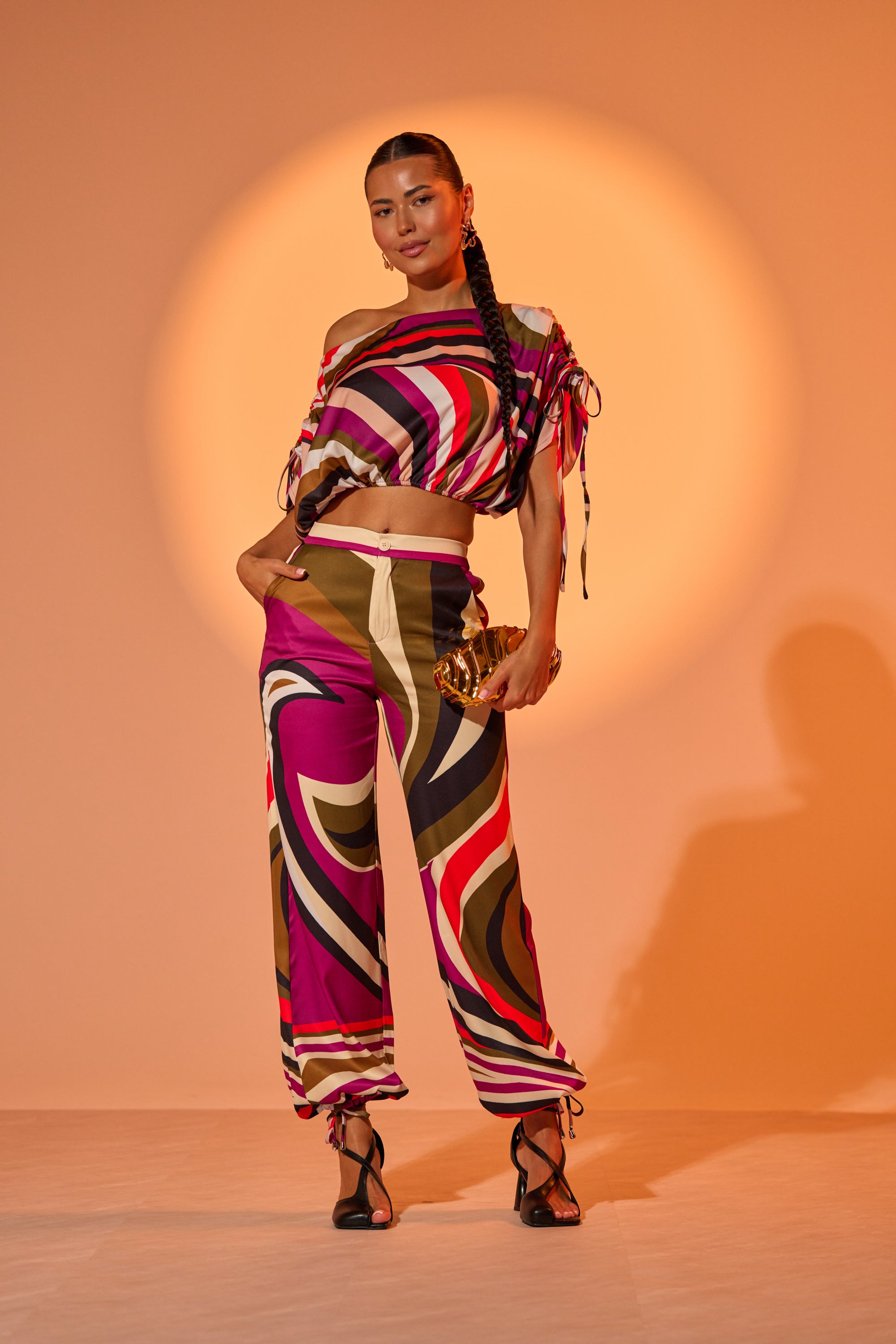 A woman stands confidently before a peach backdrop in the CRAZY IN LOVE TROUSER—a high-rise, wide-leg pant with a colorful abstract pattern—paired with a matching off-shoulder top, gold clutch, strappy heels, and a long braid. 1