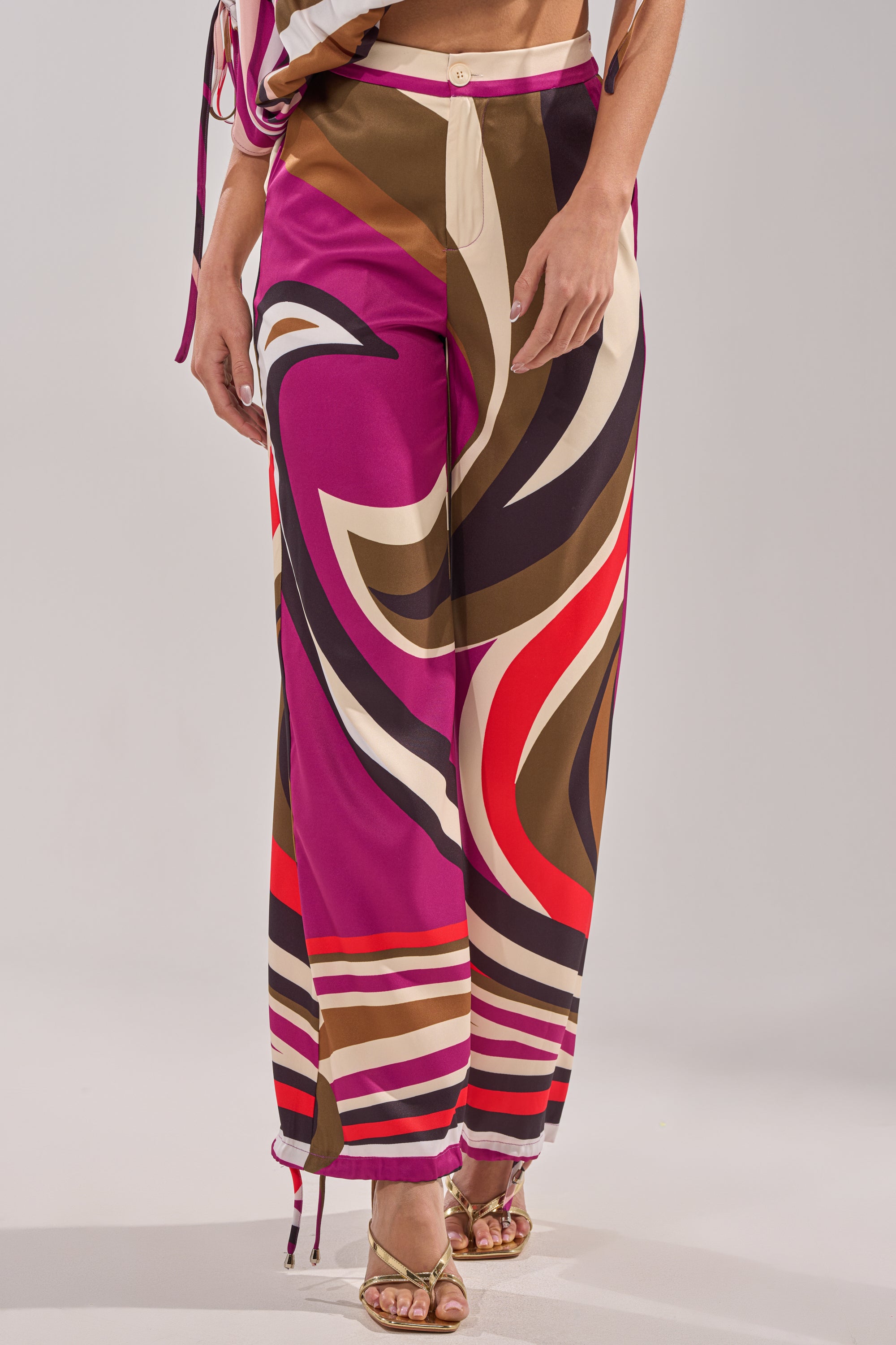 A person models the CRAZY IN LOVE TROUSER—retro-chic, high-rise, wide-leg pants with a bold abstract pattern in pink, brown, cream, red, and black—paired with open-toe heels. Upper body is partially cropped out. 3