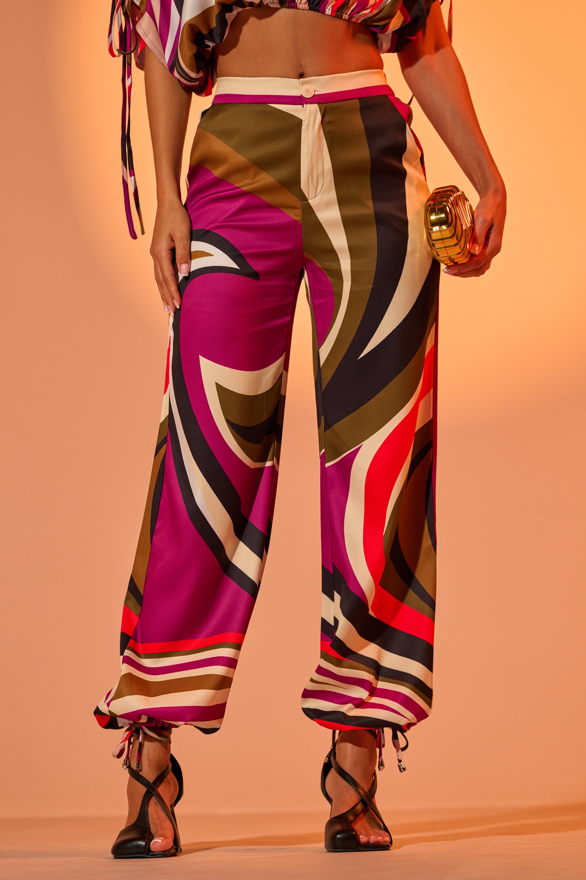 A woman wears the CRAZY IN LOVE TROUSER, a retro-chic wide-leg pant featuring a vibrant abstract swirl pattern in pink, purple, olive, and cream. She pairs it with strappy black heels and a gold clutch; upper body is partially cropped out. 0