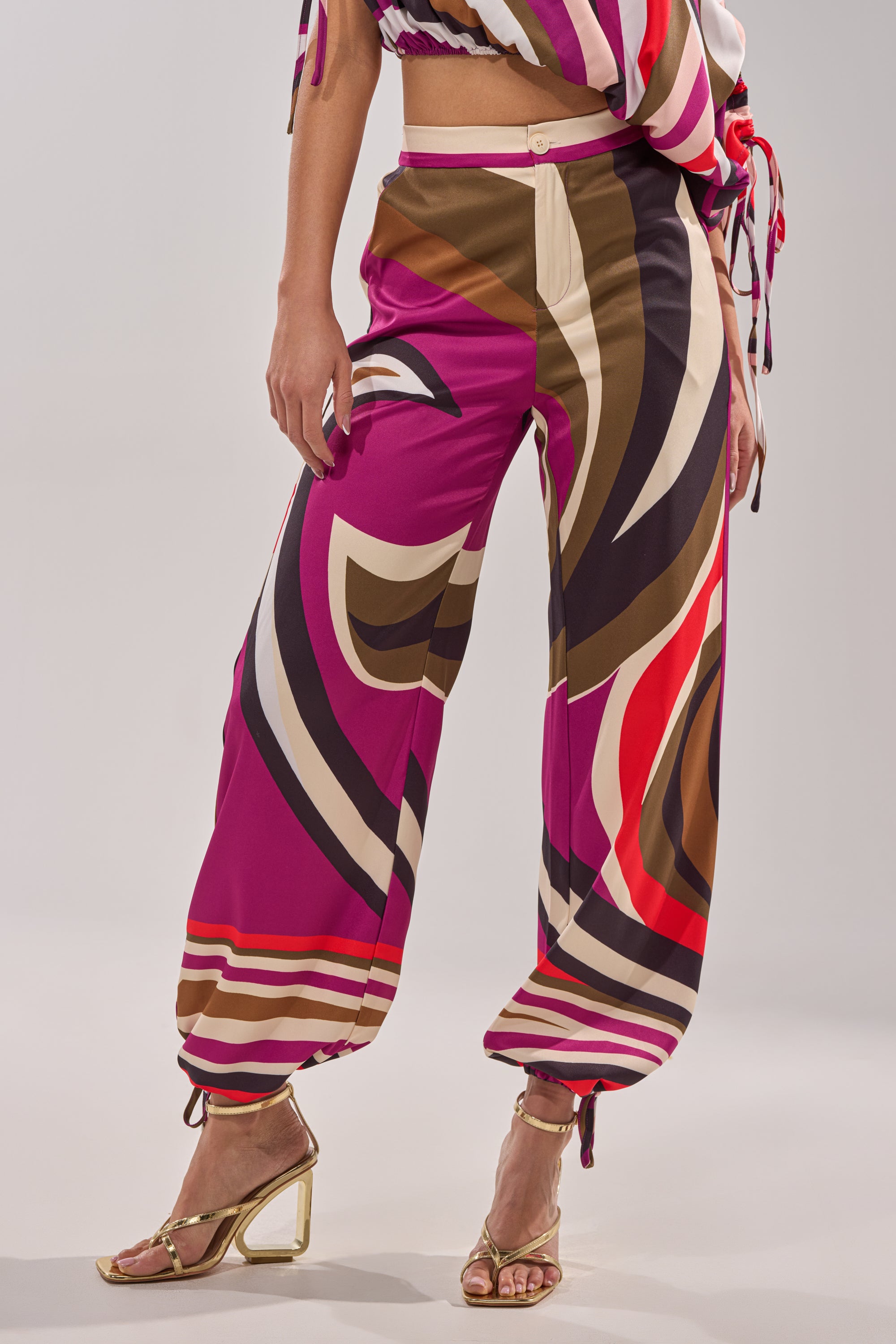 Wearing the CRAZY IN LOVE TROUSER, a retro-chic wide-leg pant with a high-rise waist and bold swirling patterns in pink, cream, brown, and red. Paired with the matching top and gold heels against a light background. 4