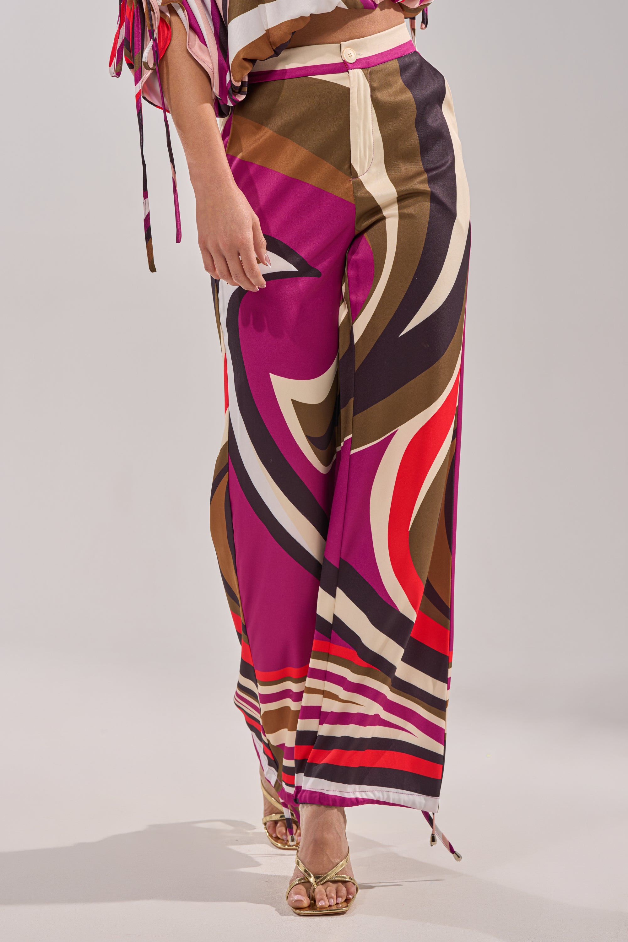 Model wearing the CRAZY IN LOVE TROUSER, featuring a retro-chic wide-leg silhouette and bold abstract pattern in pink, brown, cream, and red, paired with strappy gold sandals against a plain light gray background. 5