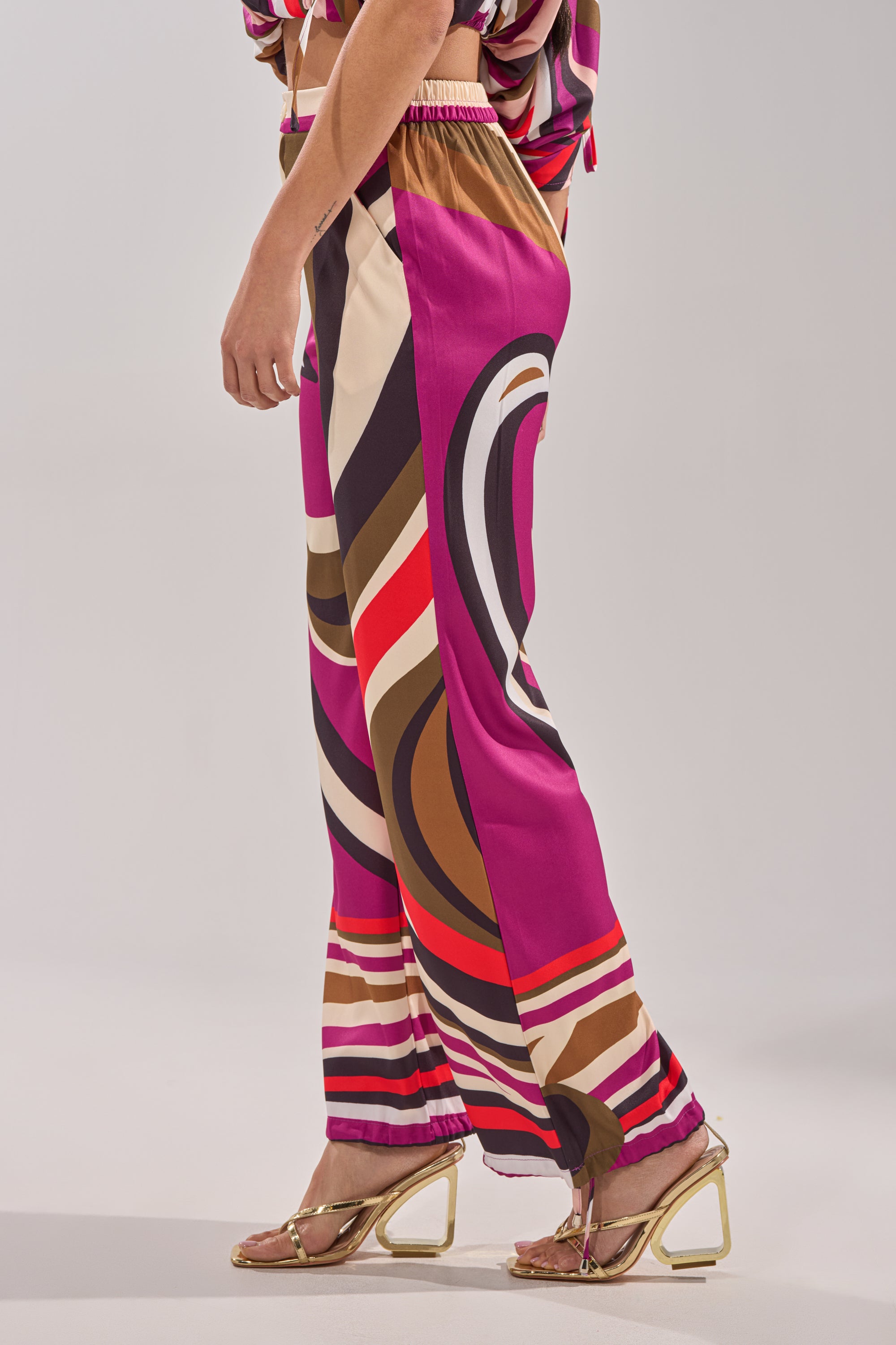 Wearing the CRAZY IN LOVE TROUSER—retro-chic, colorful wide-leg pants with a high-rise waist and bold magenta, red, black, beige, and brown swirls—paired with gold heeled sandals against a plain light background. 6
