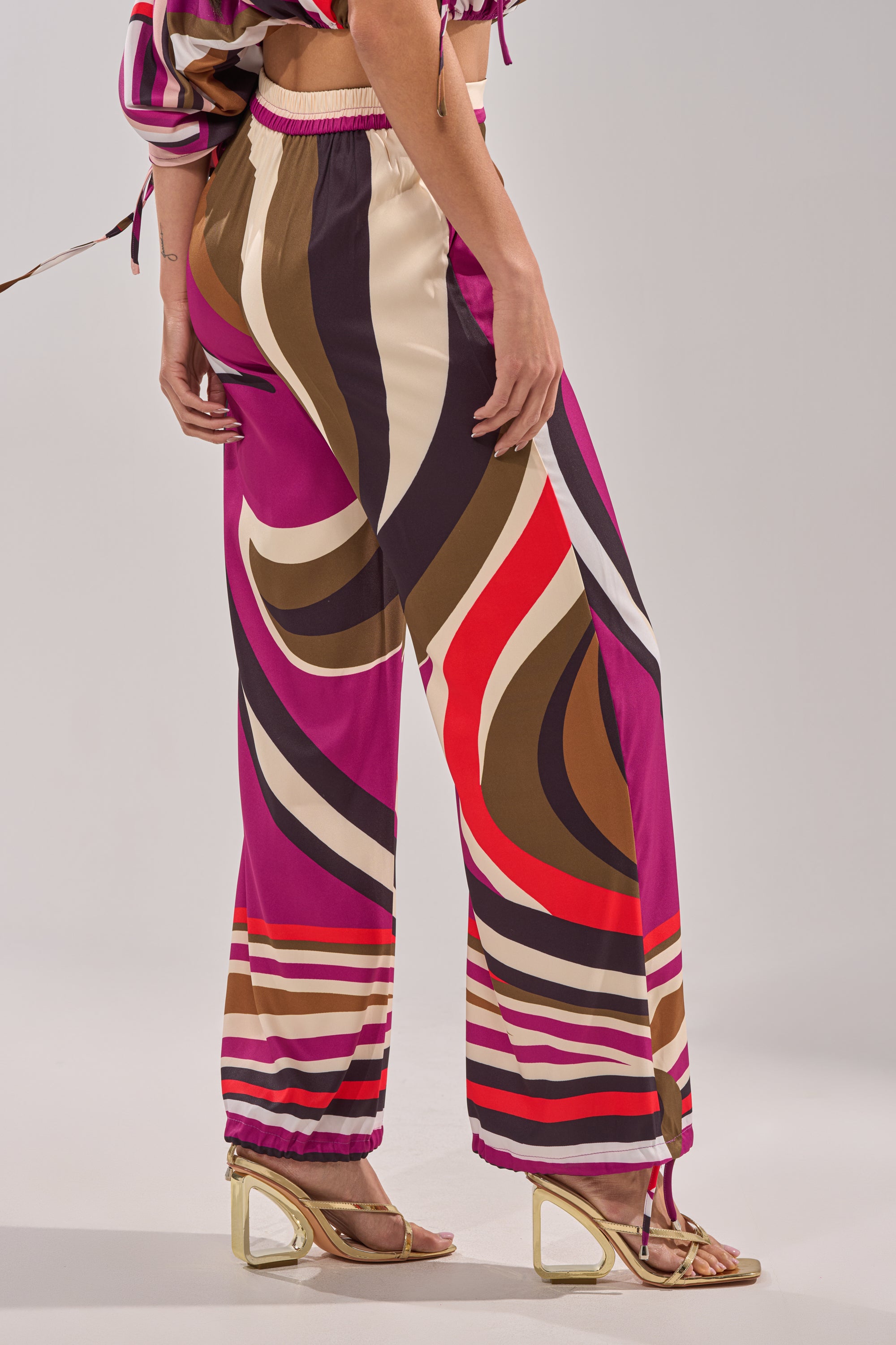 A person models the CRAZY IN LOVE TROUSER—colorful, patterned wide-leg pants with a high-rise waist—paired with open-toe heeled sandals. Only the lower half of the body is shown against a plain background. 7