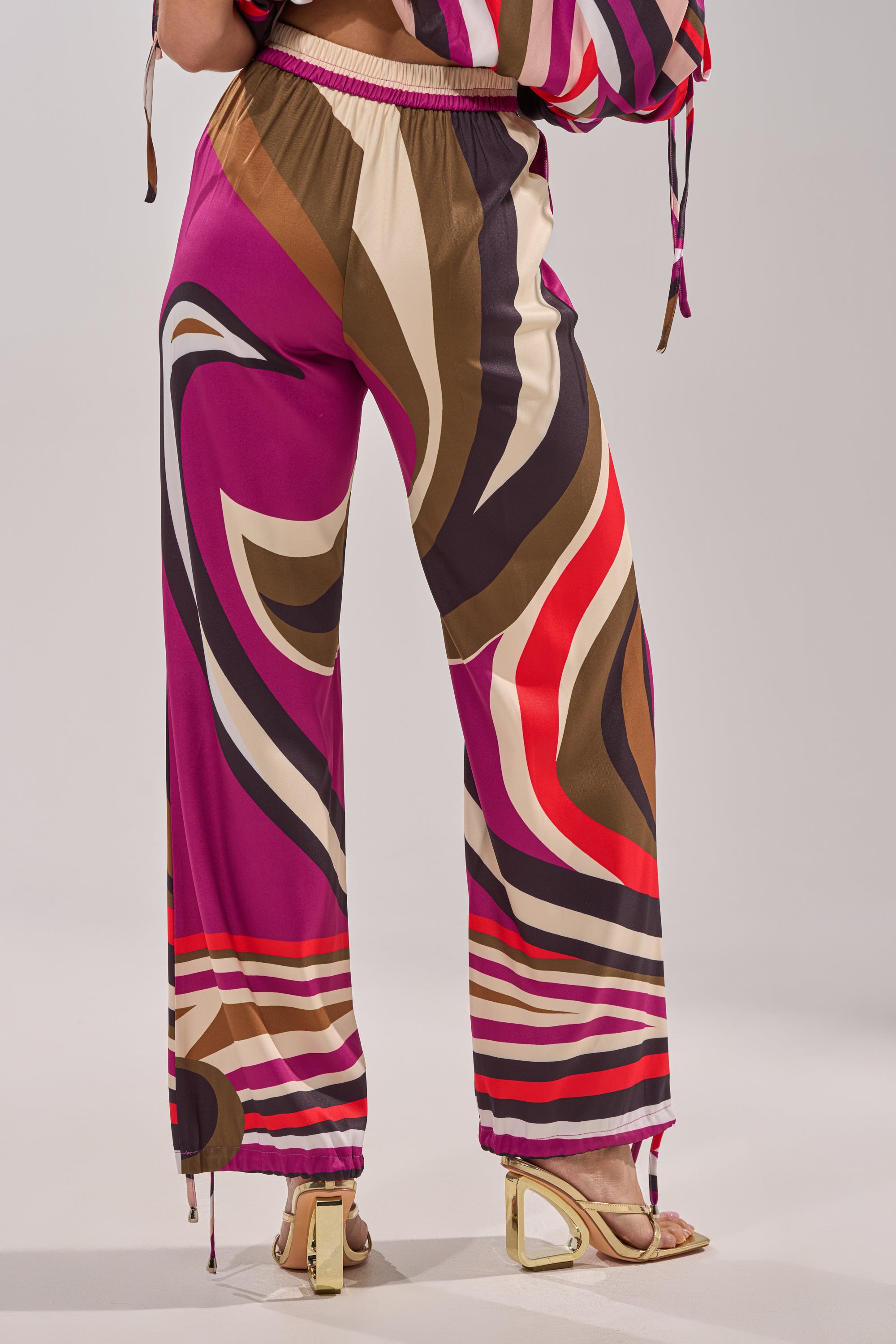 Wearing the CRAZY IN LOVE TROUSER—colorful, retro-chic wide-leg pants in pink, brown, cream, and red—a person stylishly poses with gold heeled sandals and a visible arm against a neutral background. 8