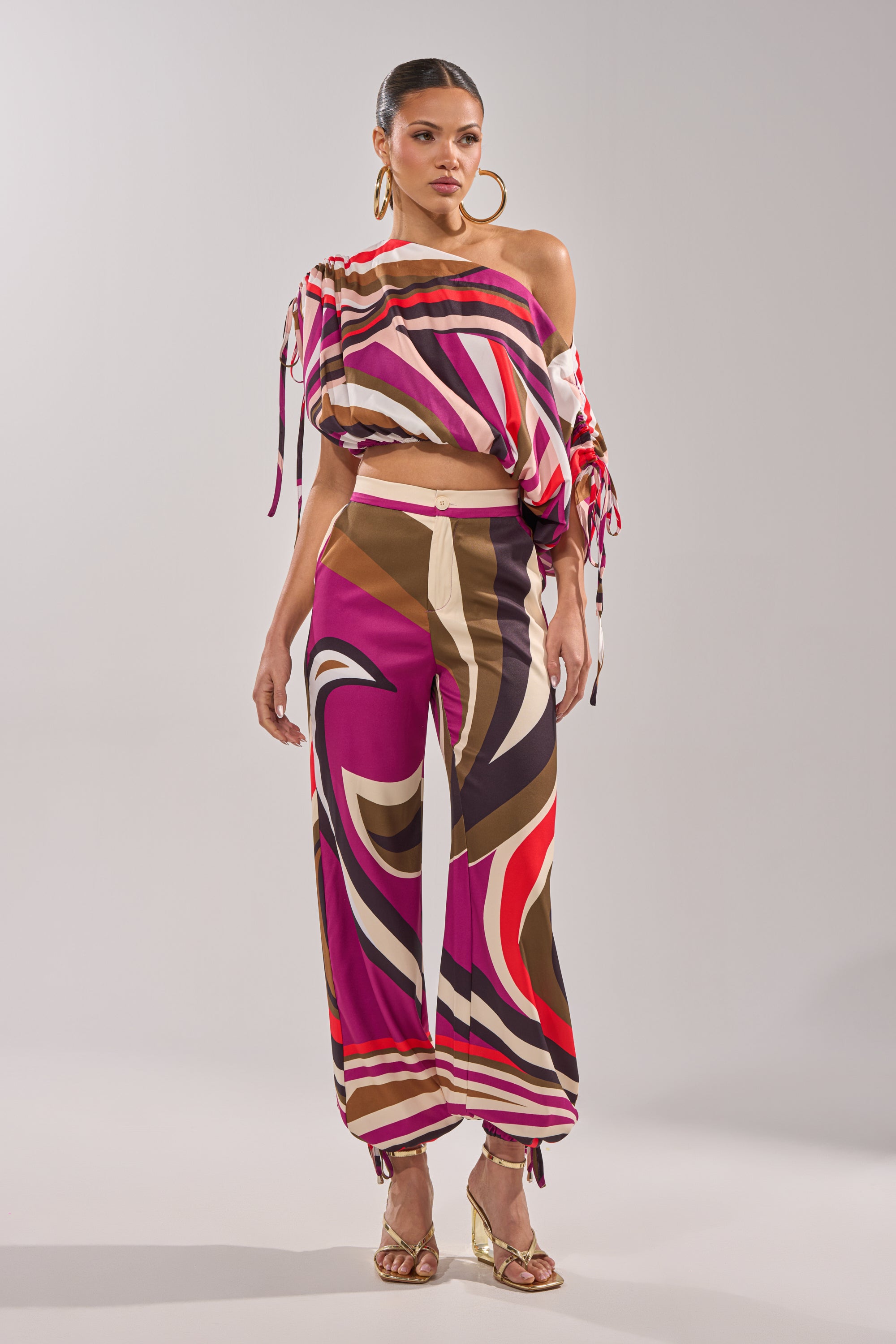 A woman poses against a plain background wearing the CRAZY IN LOVE TROUSER—a colorful, abstract-patterned wide-leg pant with a high-rise waist—paired with an off-shoulder crop top, gold hoop earrings, and gold high-heeled sandals. 9