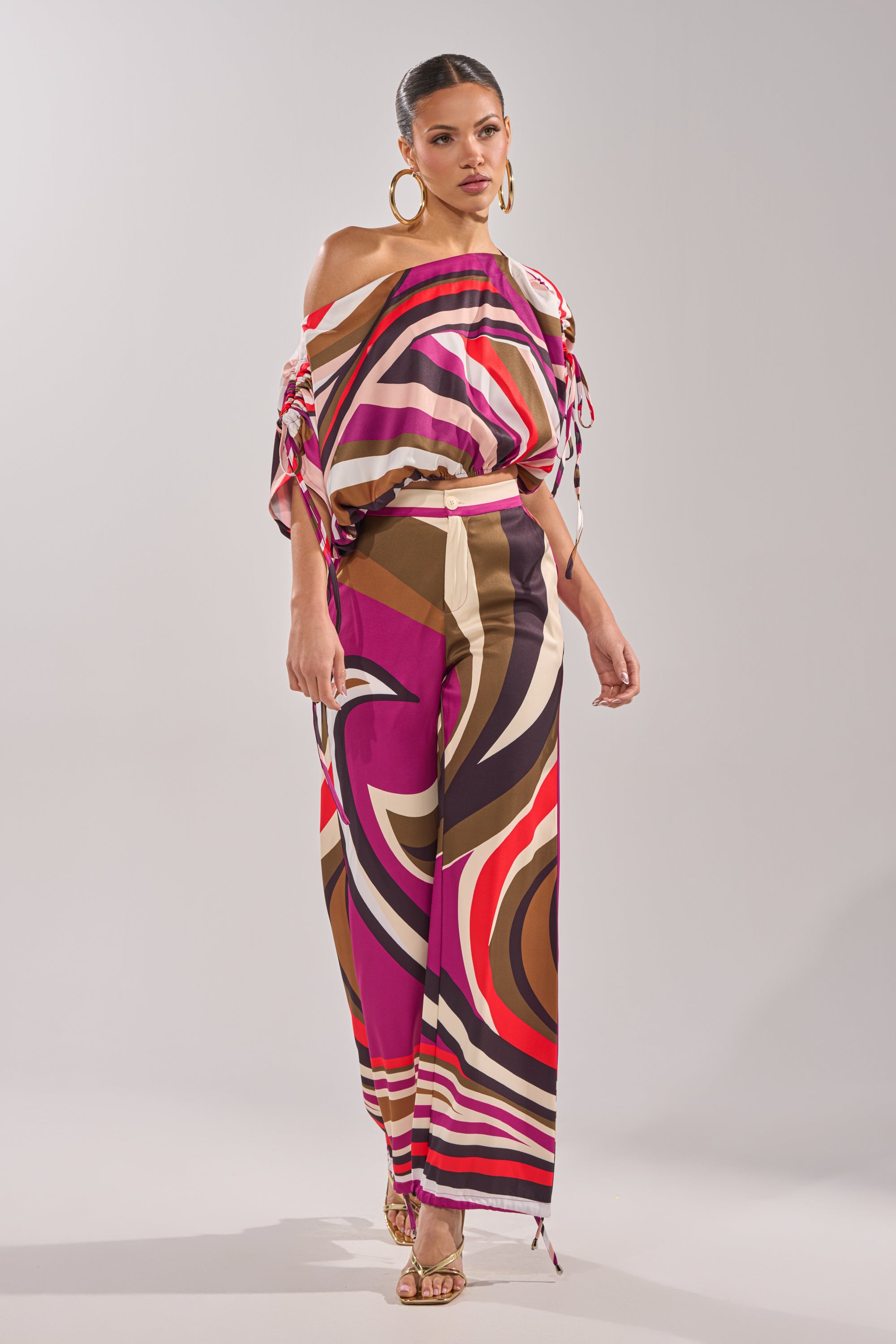A woman poses in the colorful, retro-chic CRAZY IN LOVE TROUSER featuring bold pink, red, brown, and beige swirls, paired with a loose off-shoulder top, large hoop earrings, and gold high-heeled sandals. 10