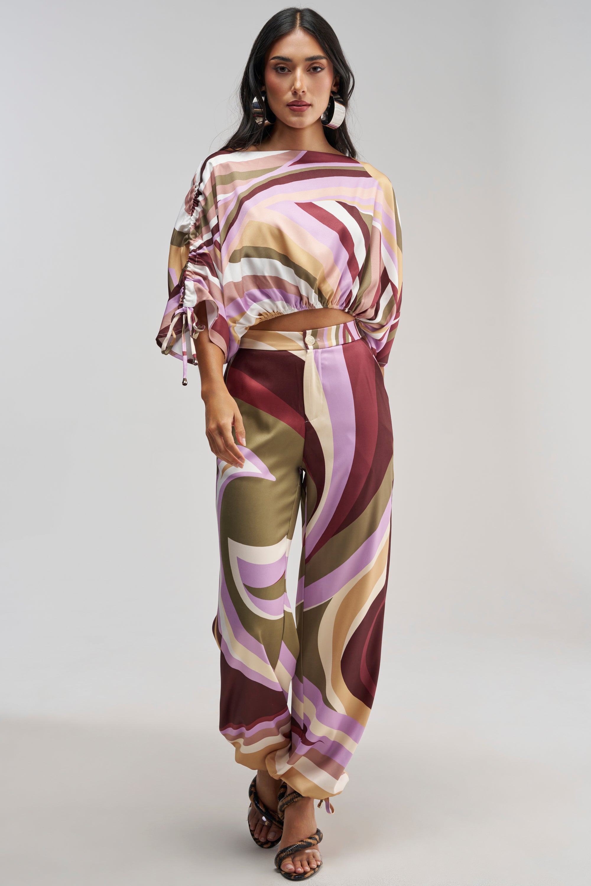 A woman is seen wearing the CRAZY FOR YOU TROUSER, featuring a high-rise waist and flowing wide-leg silhouette adorned with swirling abstract patterns in shades of maroon, pink, beige, and purple. She pairs the retro-chic pants with a loose, colorful matching top and black sandals while standing against a plain background.