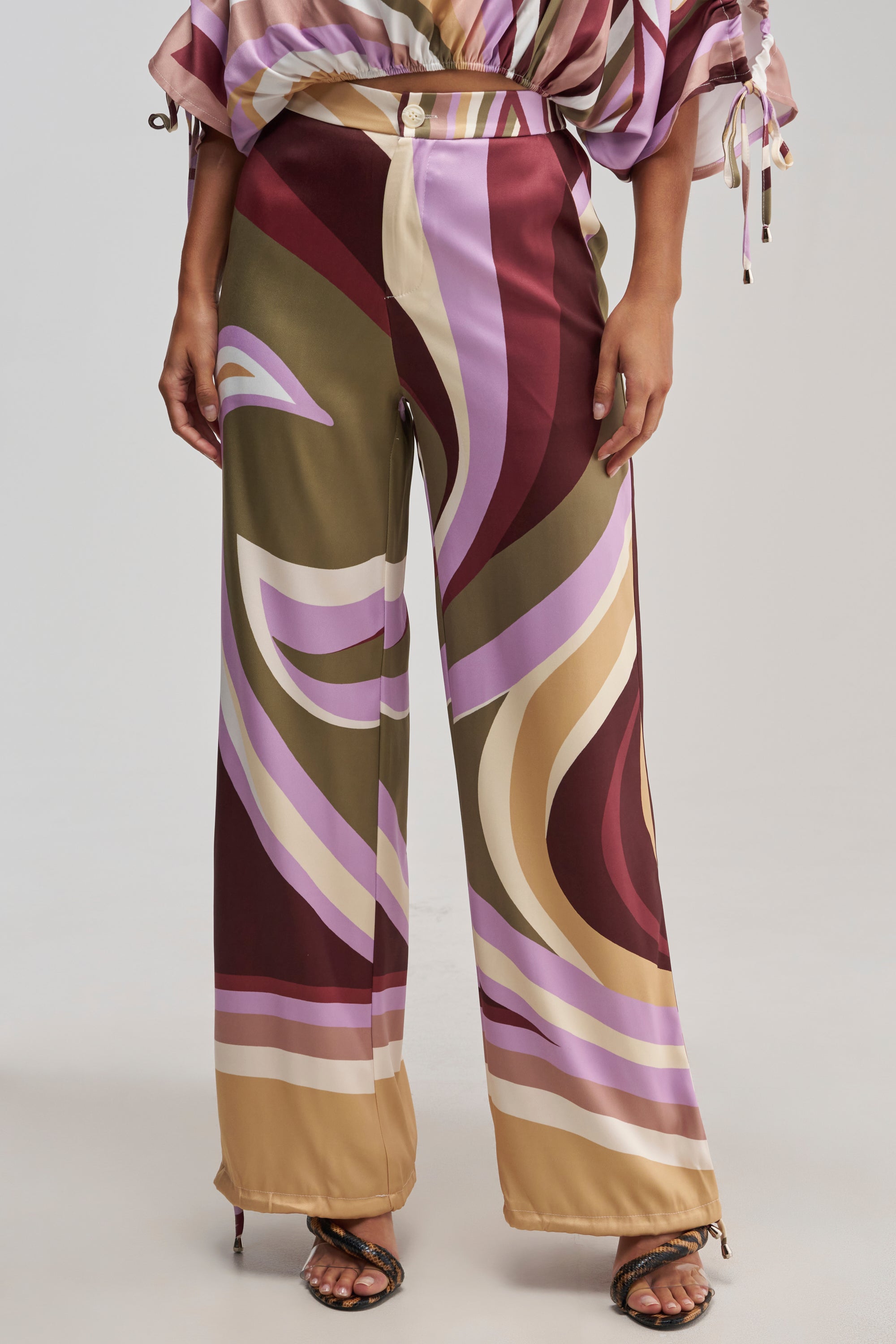 A person wearing the CRAZY FOR YOU TROUSER—abstract print wide-leg trousers with a bold swirl pattern in burgundy, olive, beige, and lavender tones and a high-rise waist—paired with open-toe sandals.