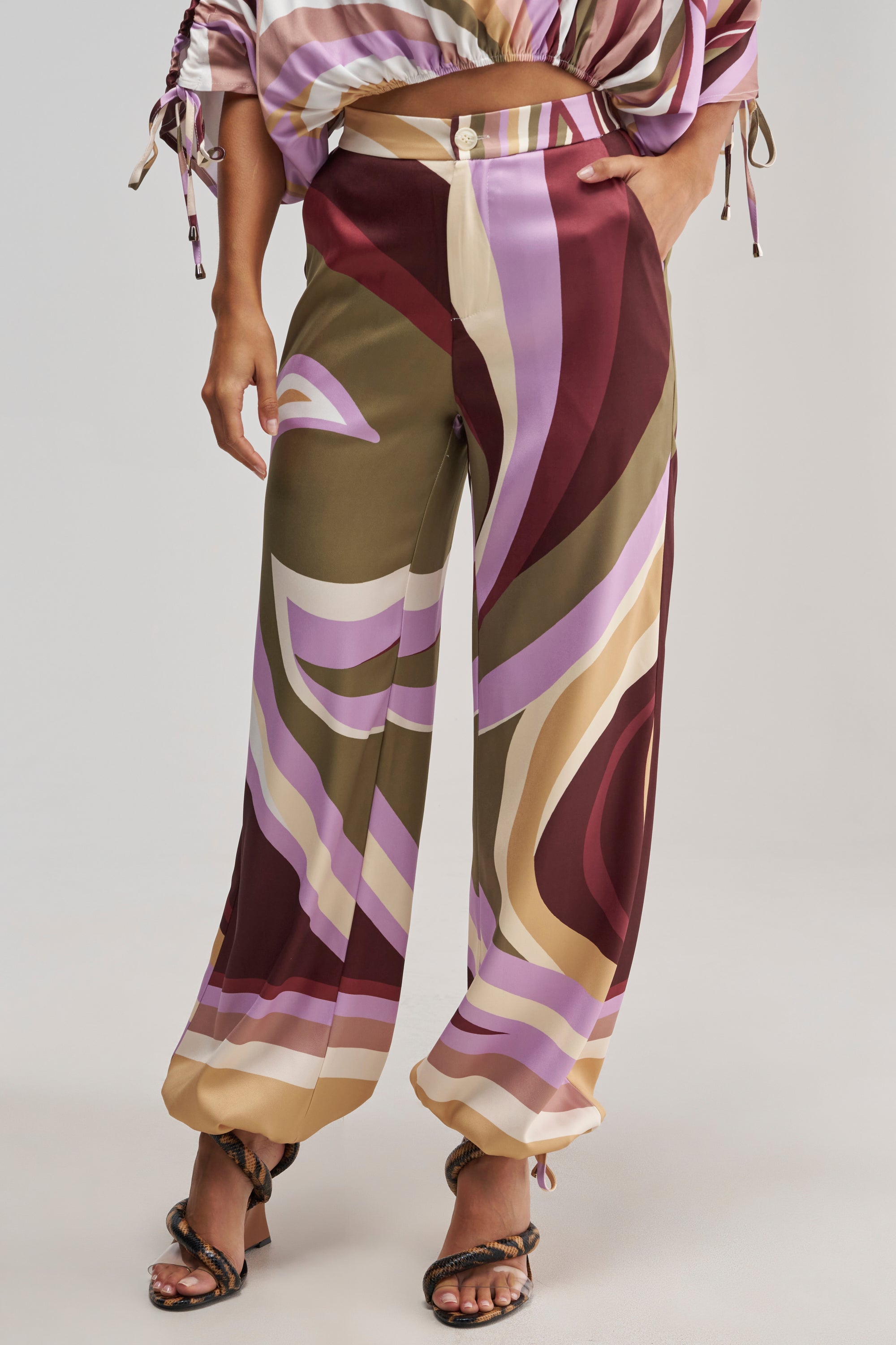 A woman wearing the CRAZY FOR YOU TROUSER, featuring a high-rise waist and wide-leg silhouette with a bold abstract print in multicolor shades of burgundy, olive, lavender, and beige; styled with strappy heels. Her matching top is partially visible.